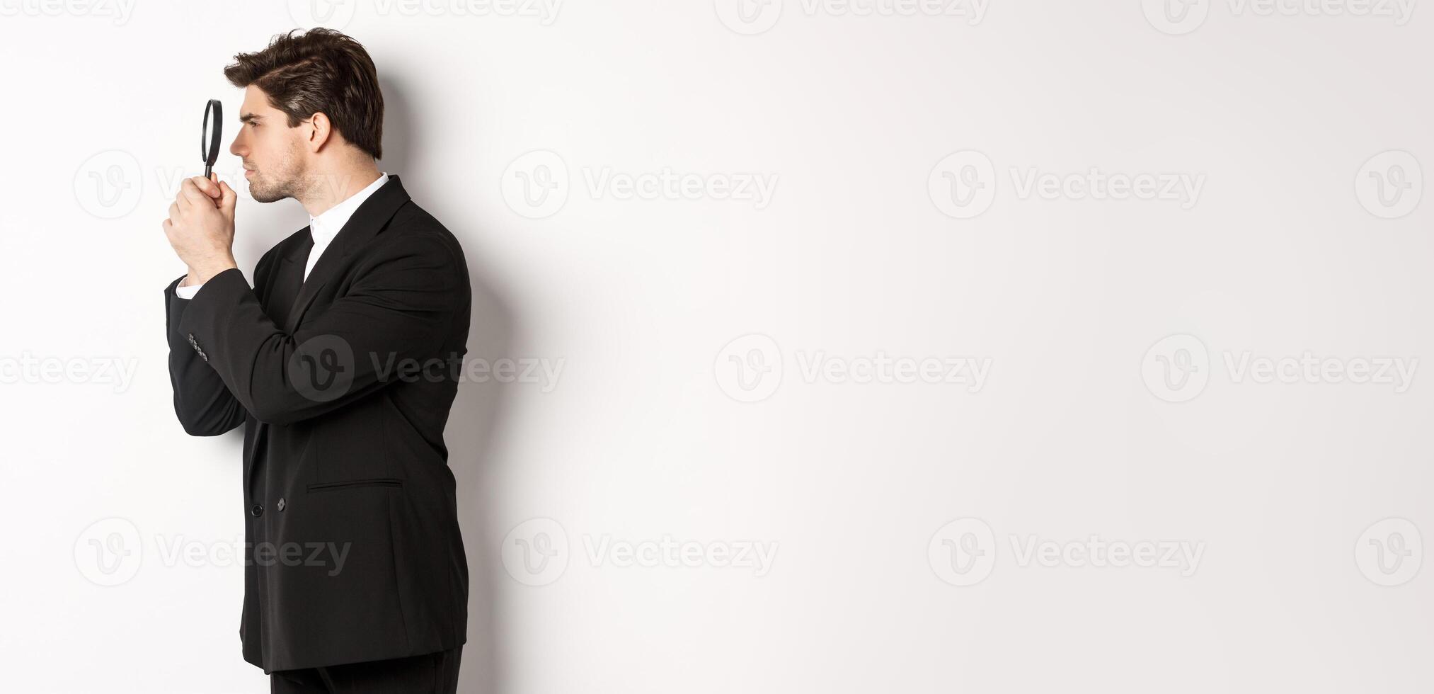 Profile shot of handsome businessman in black suit, looking through magnifying glass and searching for something, standing over white background photo