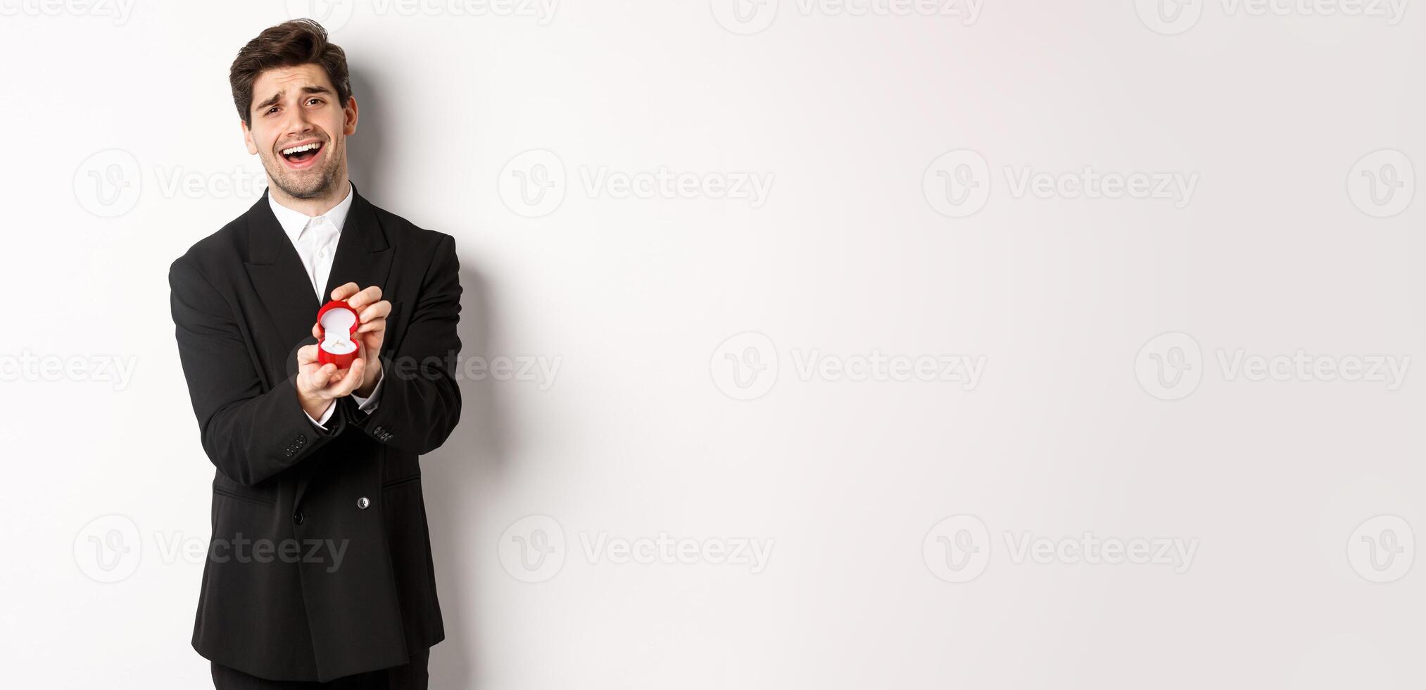 Portrait of handsome man in black suit, open box with wedding ring, making a proposal, asking to marry him, standing against white background photo