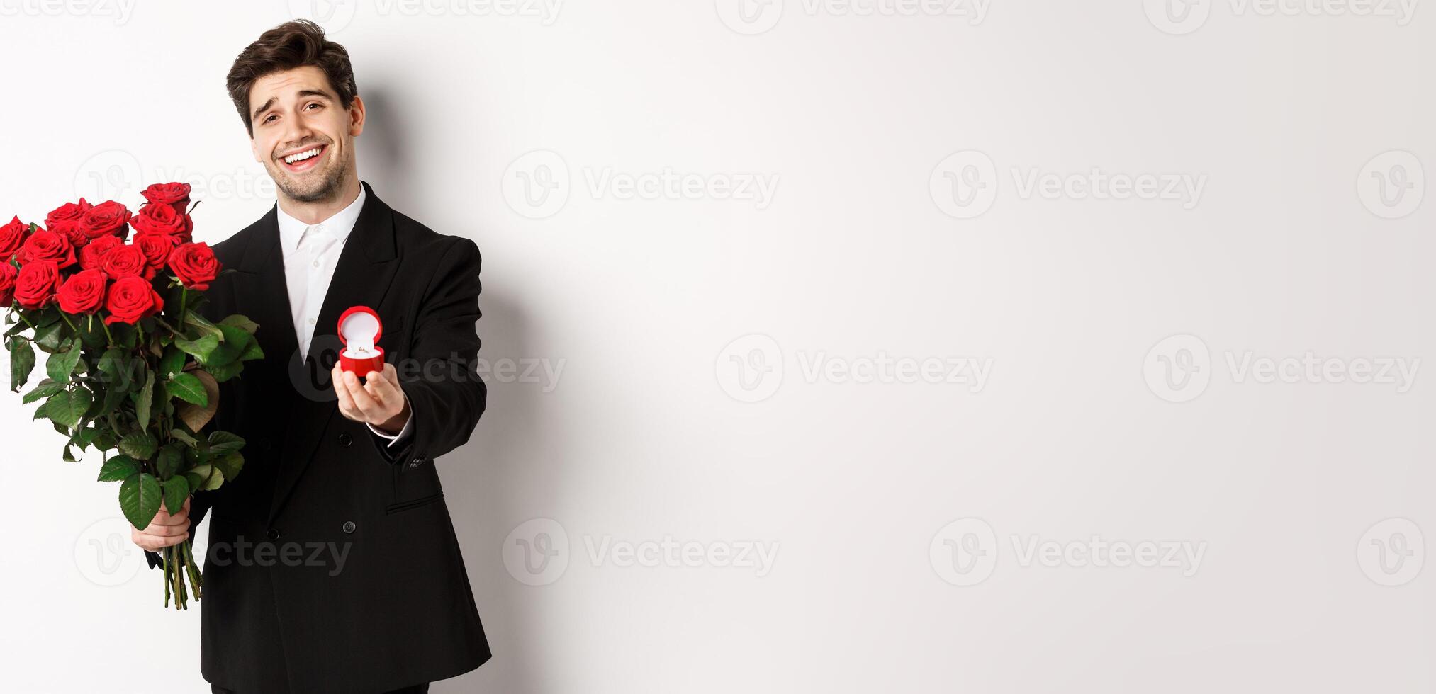 Handsome smiling man in black suit, holding roses and engagement ring, making a proposal to marry him, standing against white background photo