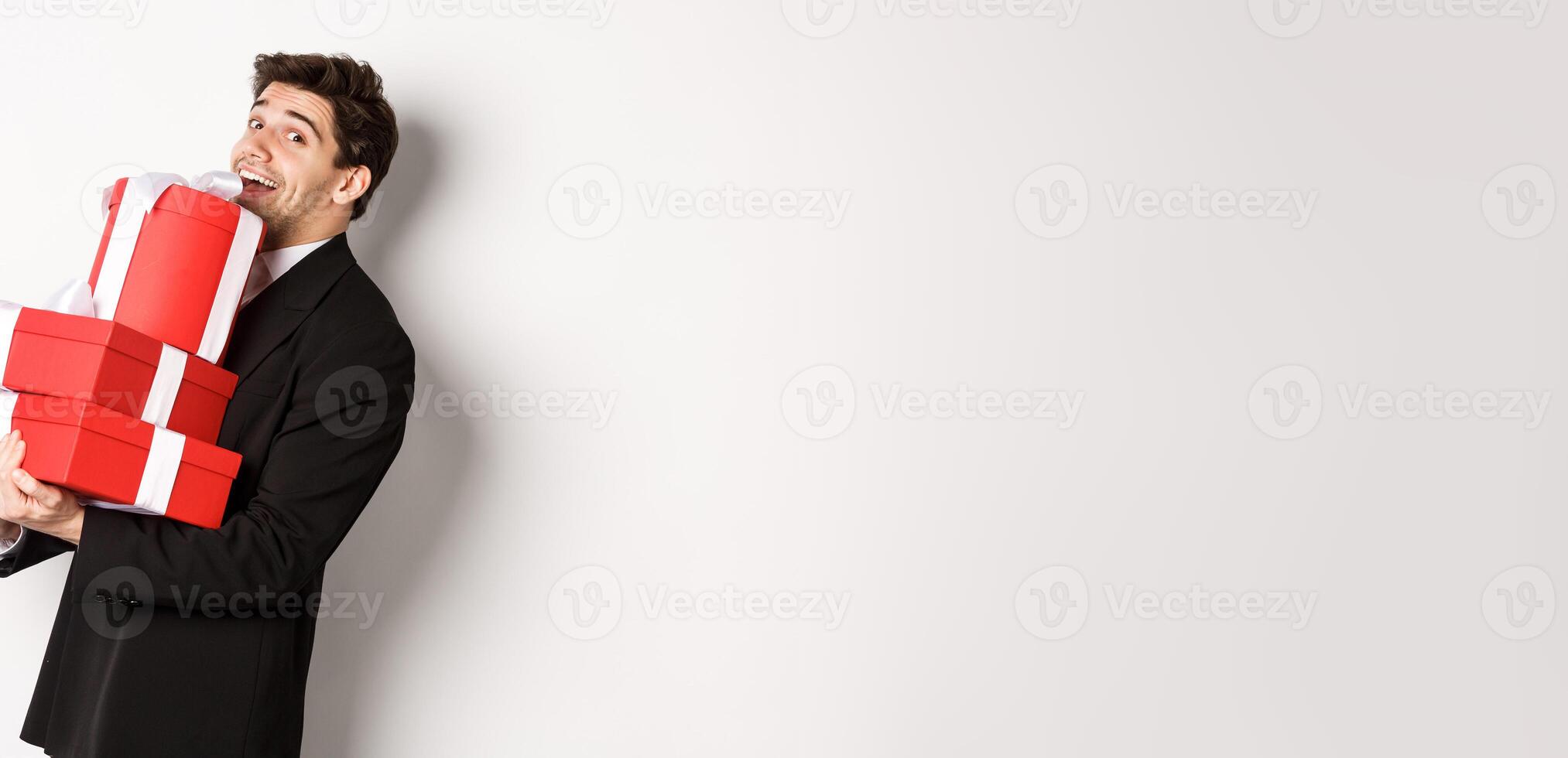 Concept of christmas holidays, celebration and lifestyle. Handsome man in black suit, holding pile of gifts and smiling, wishing happy new year, standing over white background photo