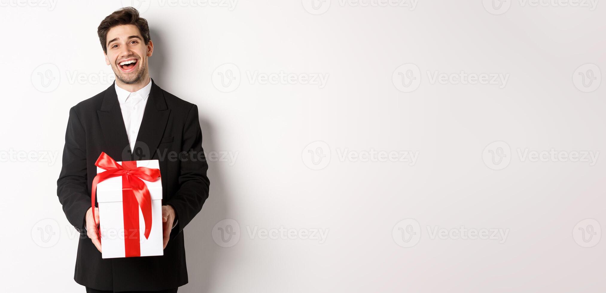 Concept of christmas holidays, celebration and lifestyle. Joyful handsome man in black suit, holding xmas gift and smiling, standing against white background photo