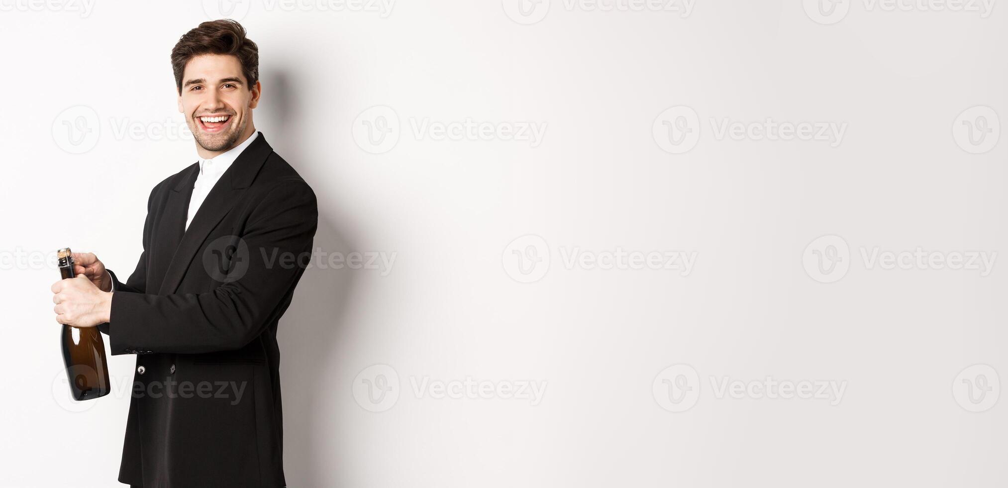 Handsome smiling man in trendy suit open a bottle of champagne, celebrating holidays, standing over white background photo