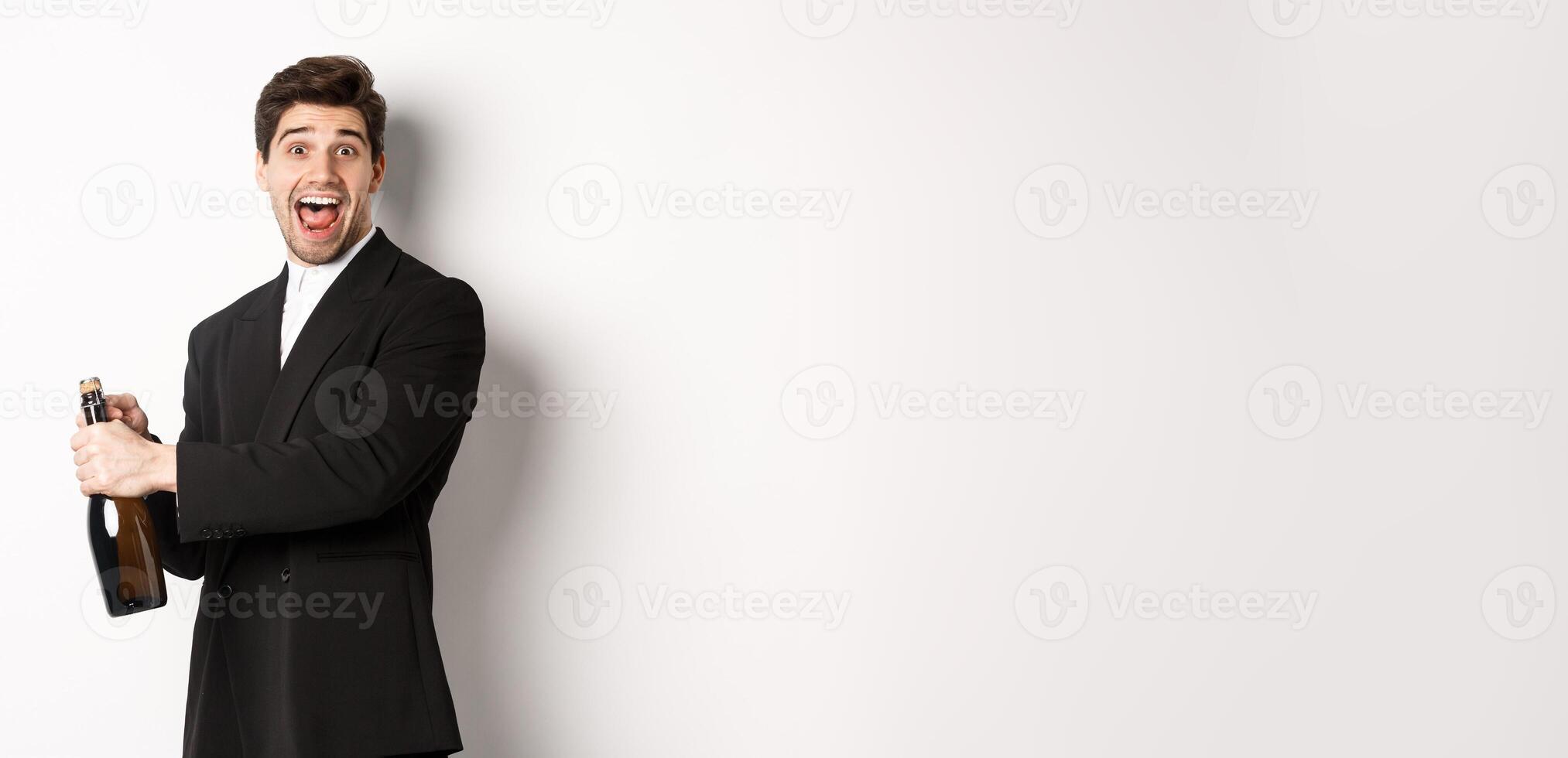 Portrait of attractive man in black suit, winking at camera and opening bottle of champagne, celebrating new year, standing against white background photo