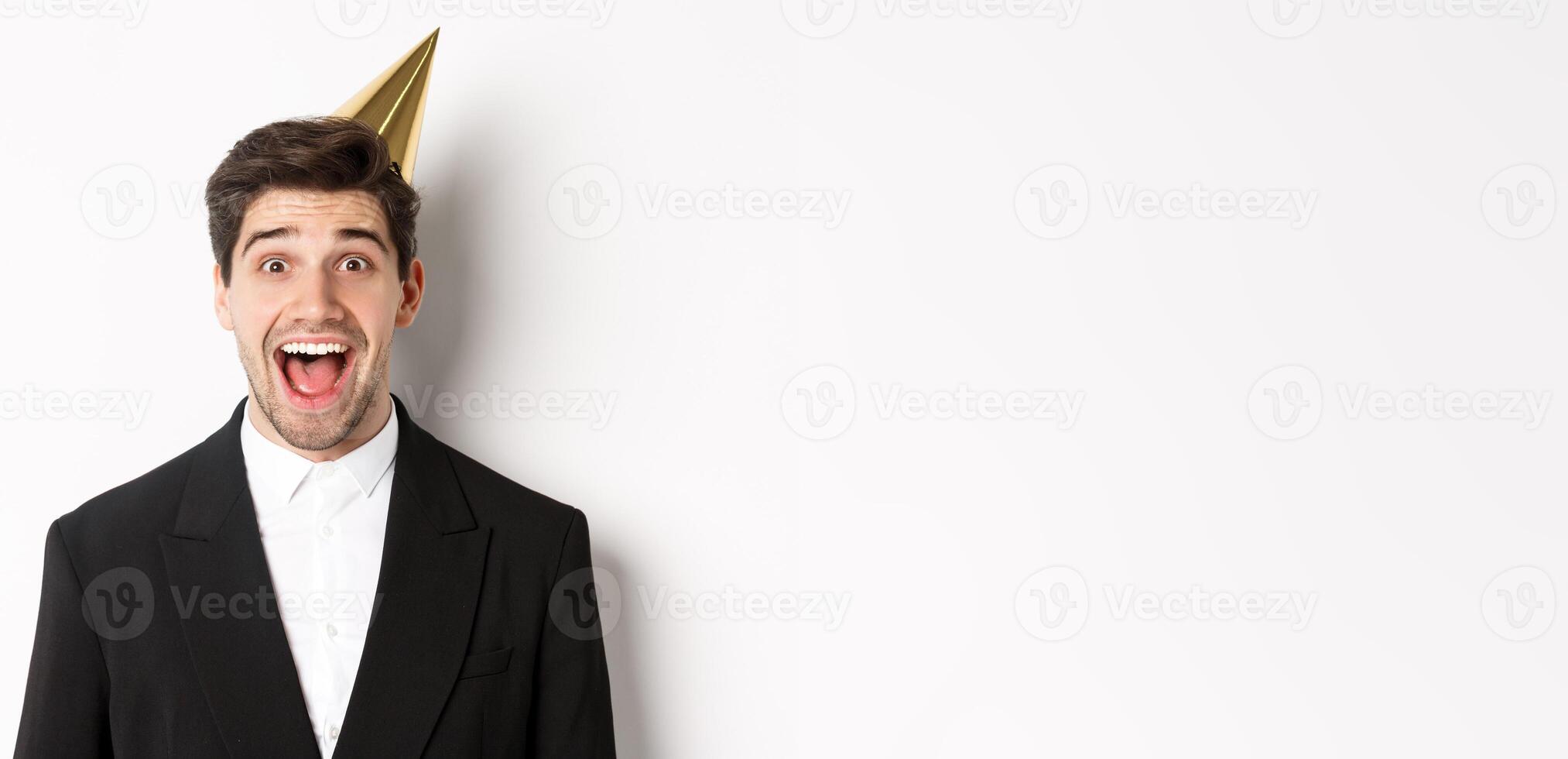 Close-up of excited handsome guy in trendy suit and party hat, looking amazed, celebrating winter holidays, standing against white background photo