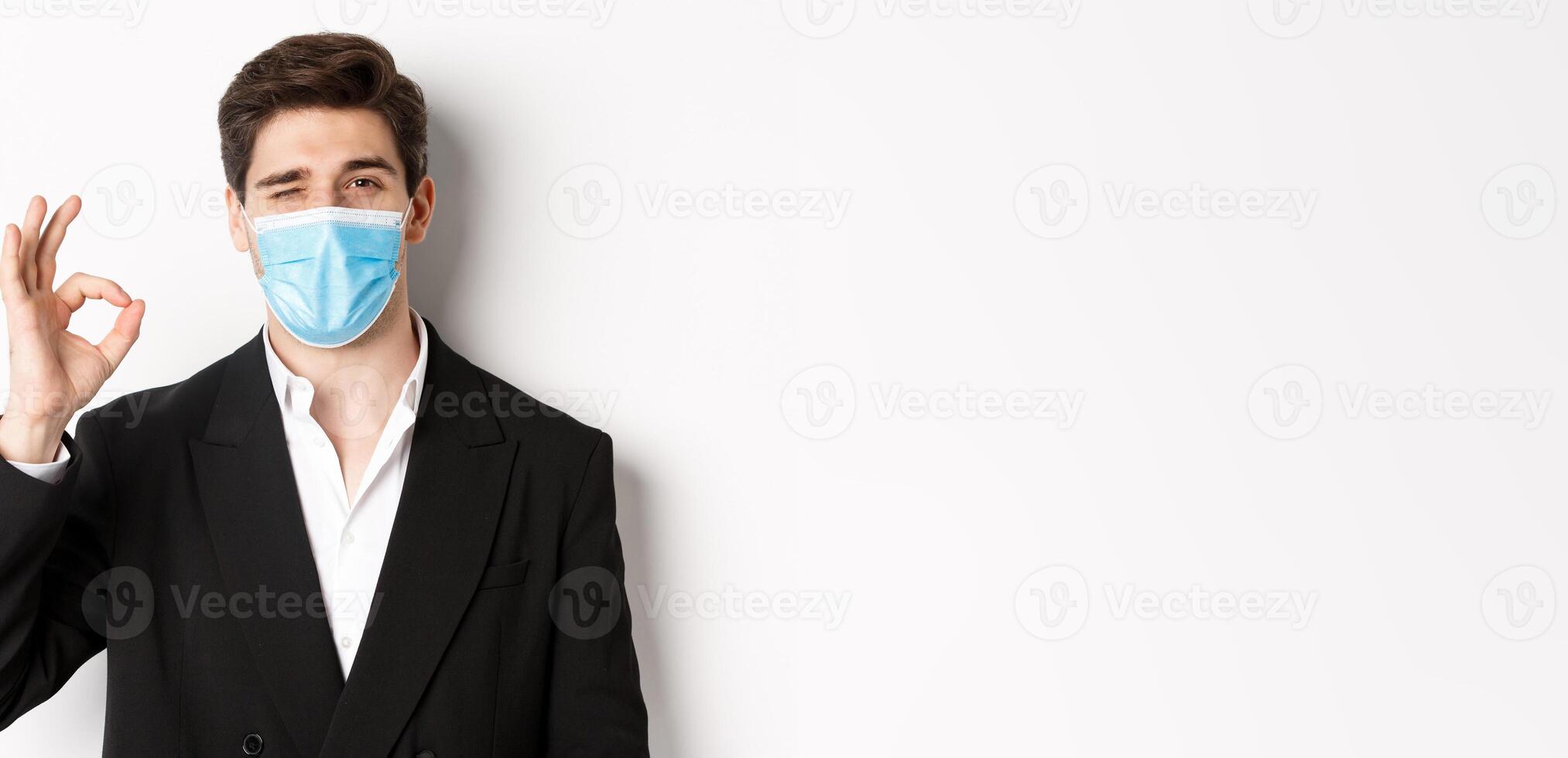 Concept of covid-19, business and social distancing. Close-up of good-looking businessman in trendy suit and medical mask, showing okay sign and winking, standing against white background photo