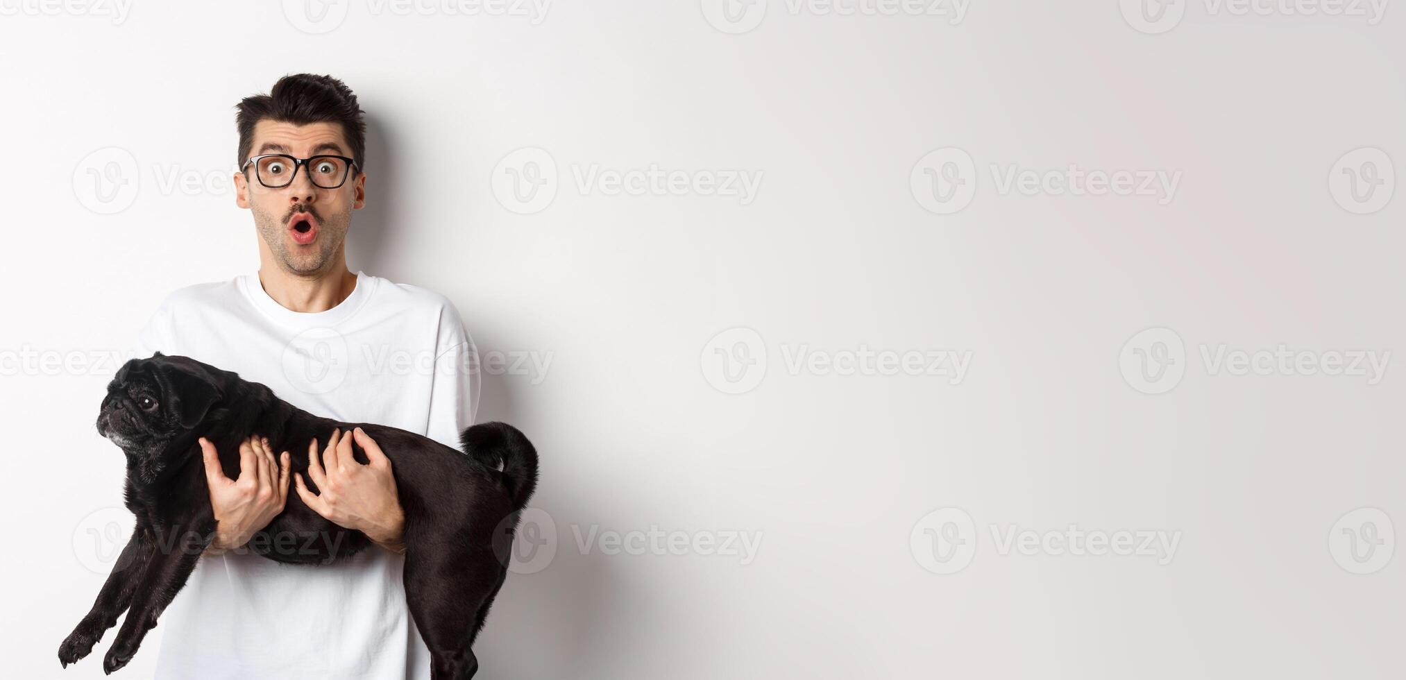 Surprised young man in glasses holding cute black pug, dog owner staring at camera with impressed face, saying wow, standing over white background photo