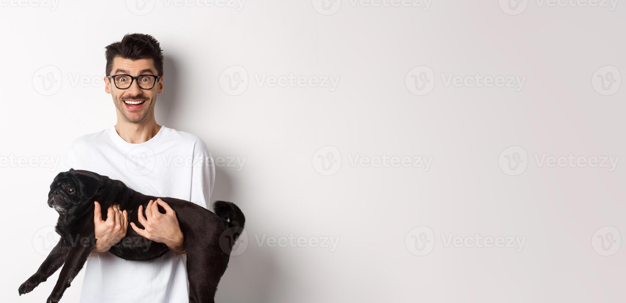 Happy young man in glasses holding black pug pet and staring at camera amused, smiling cheerful, standing over white background photo