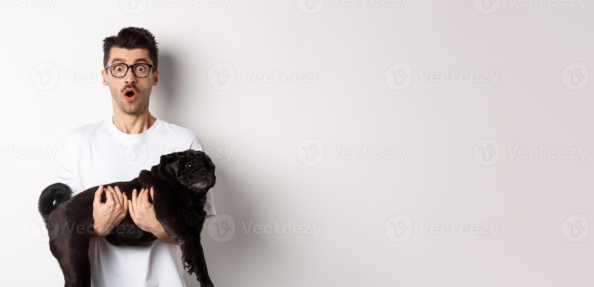 Surprised young man in glasses holding cute black pug, dog owner staring at camera with impressed face, saying wow, standing over white background photo