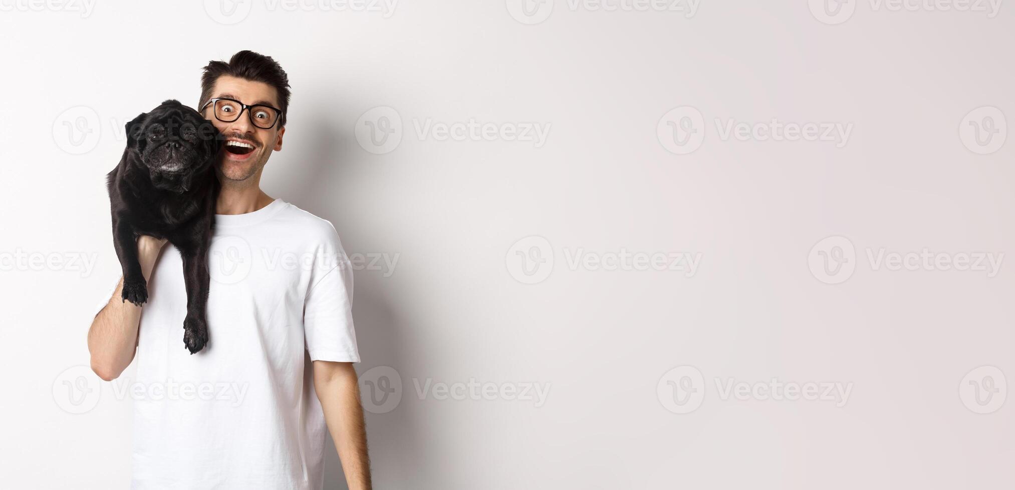 Cheerful hipster guy in glasses and t-shirt, carry cute black pug on shoulder and smiling. Dog owner playing with his pet, standing over white background photo