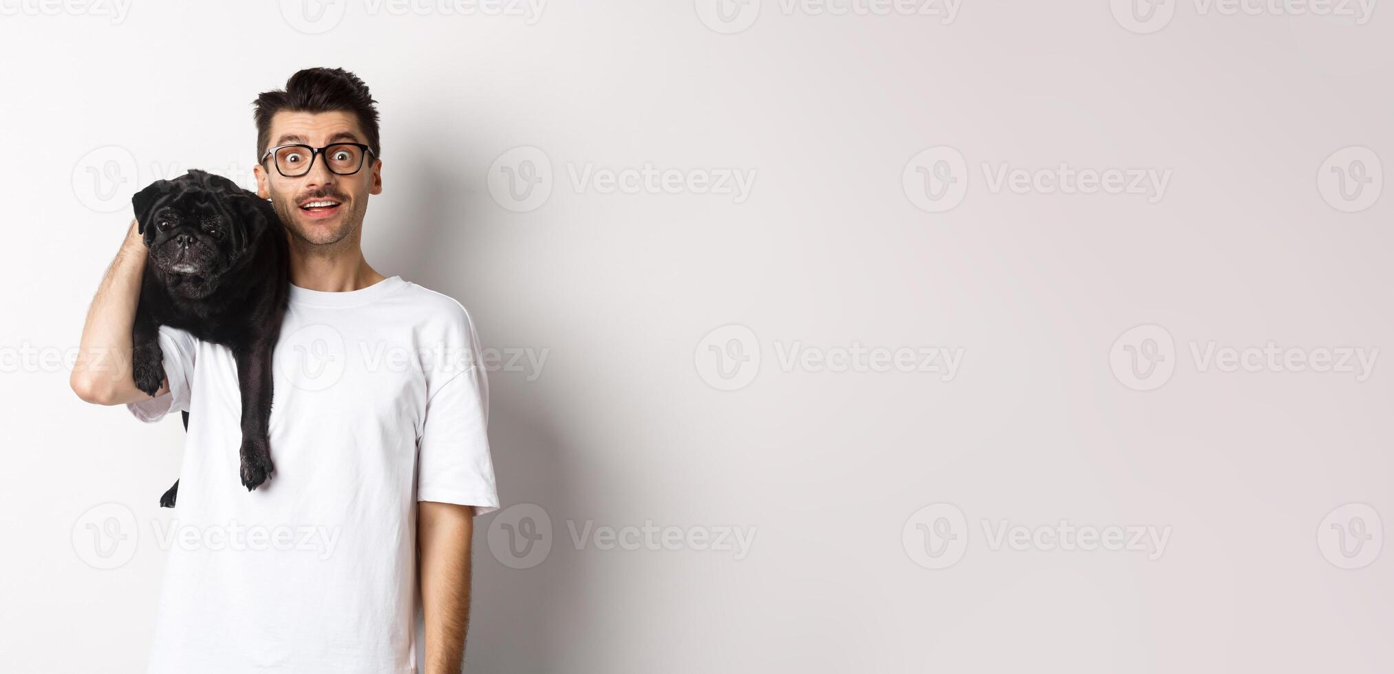 Amazed young man in glasses holding black pug on shoulder and staring at camera impressed. Dog owner posing with cute puppy near white background photo