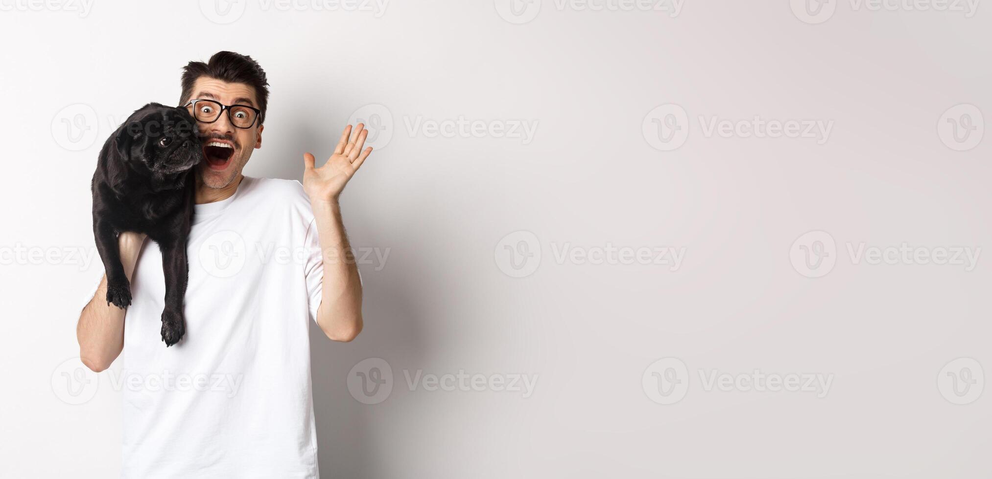 Handsome young man in glasses holding his black pug and waving hand, guy saying hello while carry dog with one arm, standing over white background photo