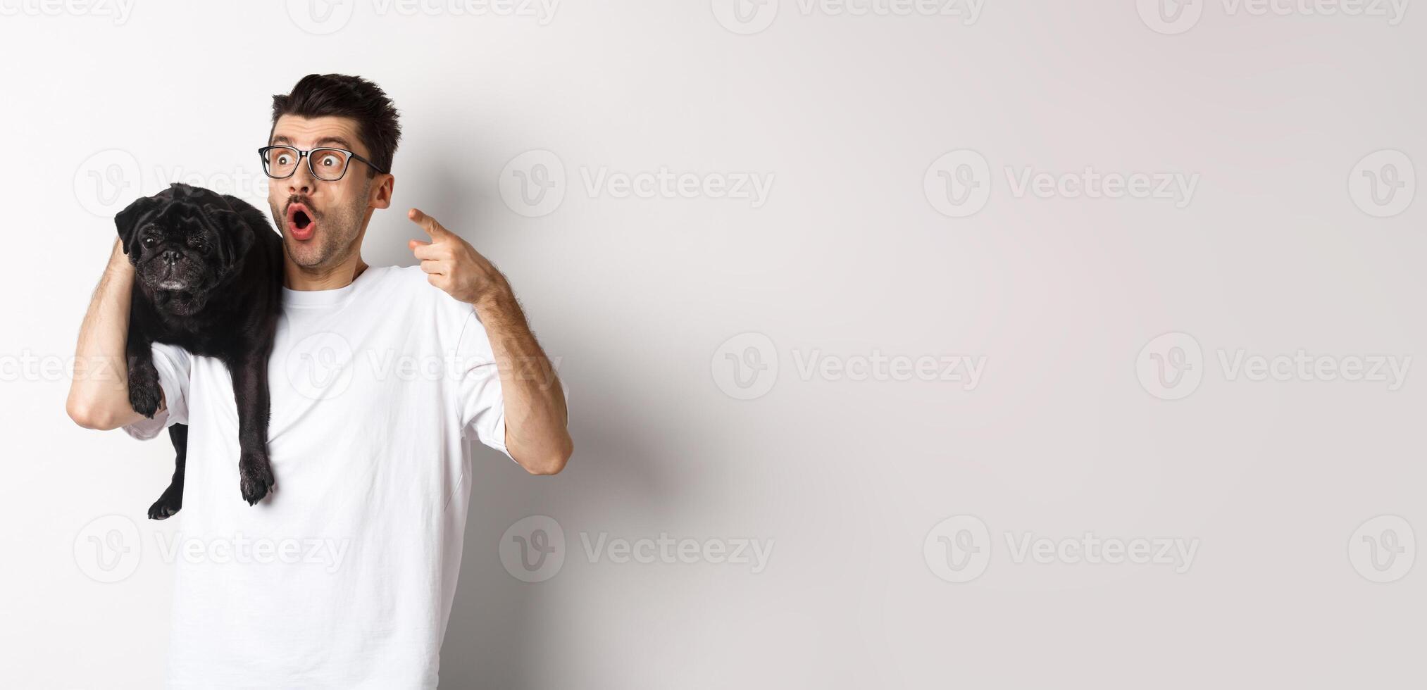 Amazed young man holding cute black dog on shoulder, pointing finger left at promo offer, staring impressed and speechless, standing over white background photo