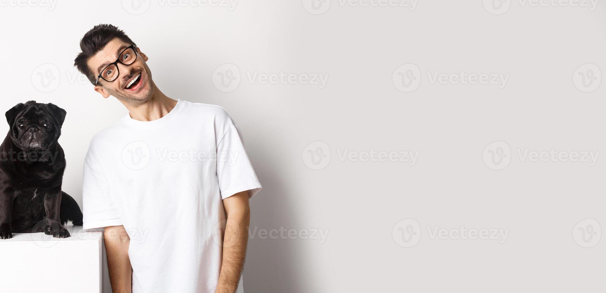 Image of handsome young man standing near cute black pug and smiling. Dog owner spending time with his pet, staring at camera happy, white background photo
