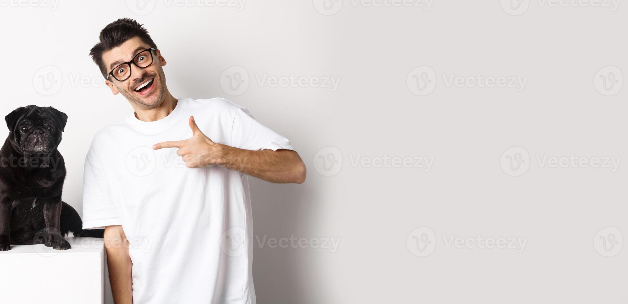 Happy young man showing his cute dog, pointing finger at black pug and smiling, standing over white background photo