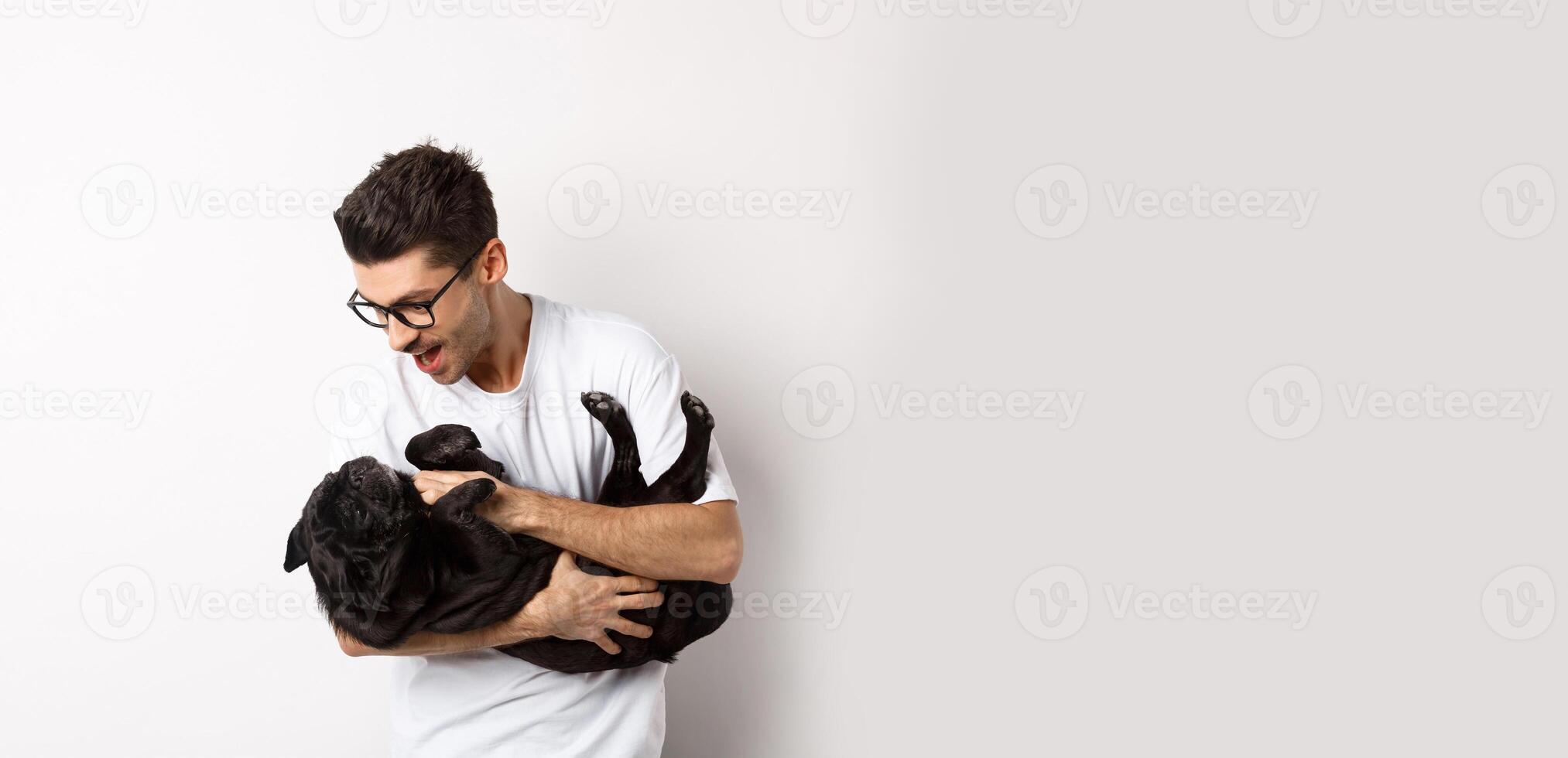 Handsome young man playing with cute black puppy. Dog owner petting a pug, standing over white background photo