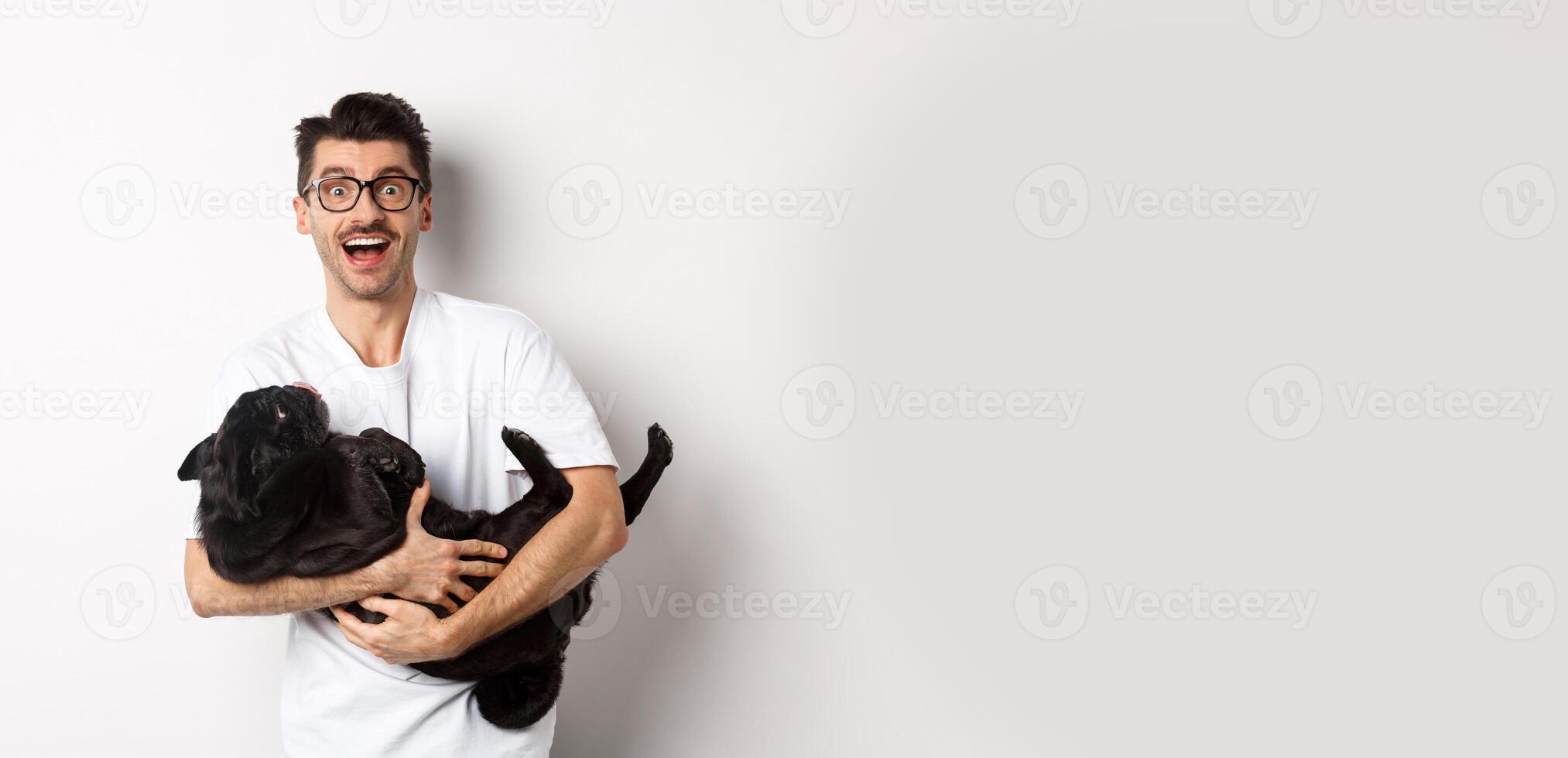 Handsome young man playing with cute black puppy. Dog owner petting a pug, standing over white background photo
