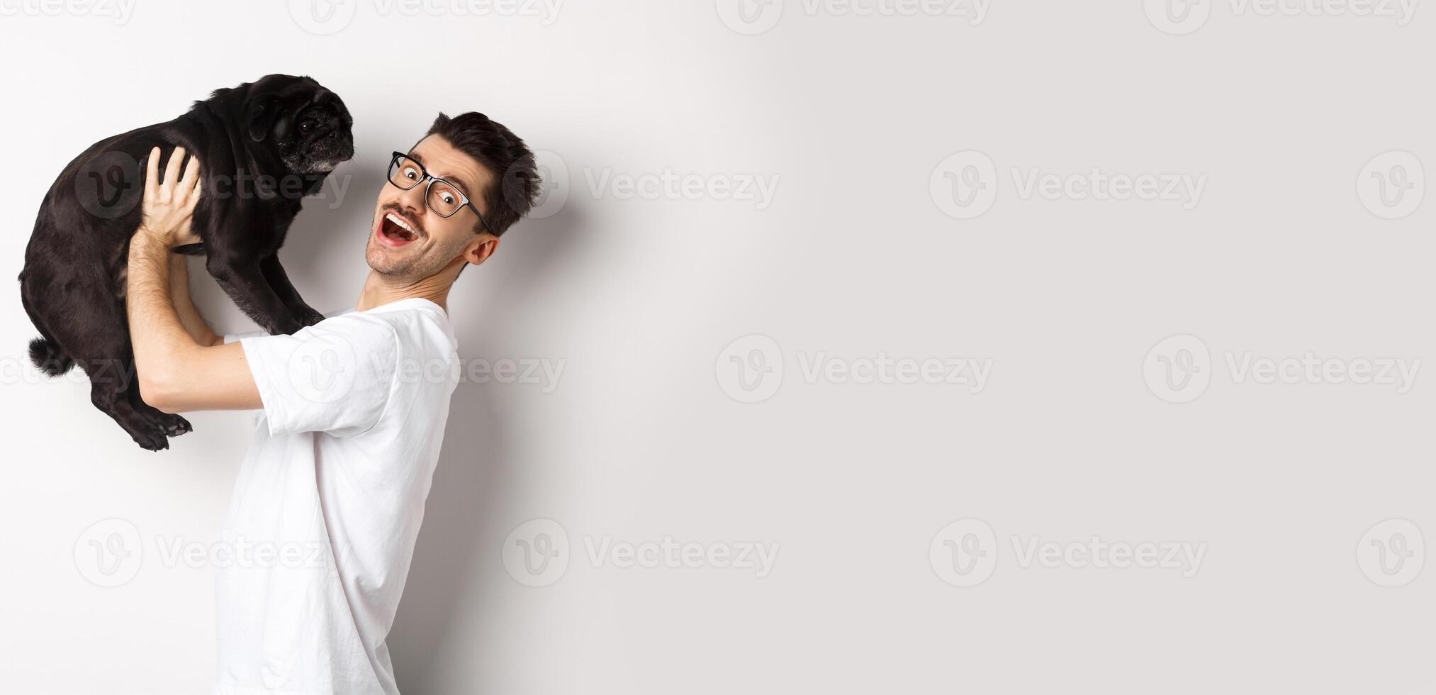 Image of handsome young man loving his pug. Dog owner holding puppy and smiling happy at camera, standing over white background photo