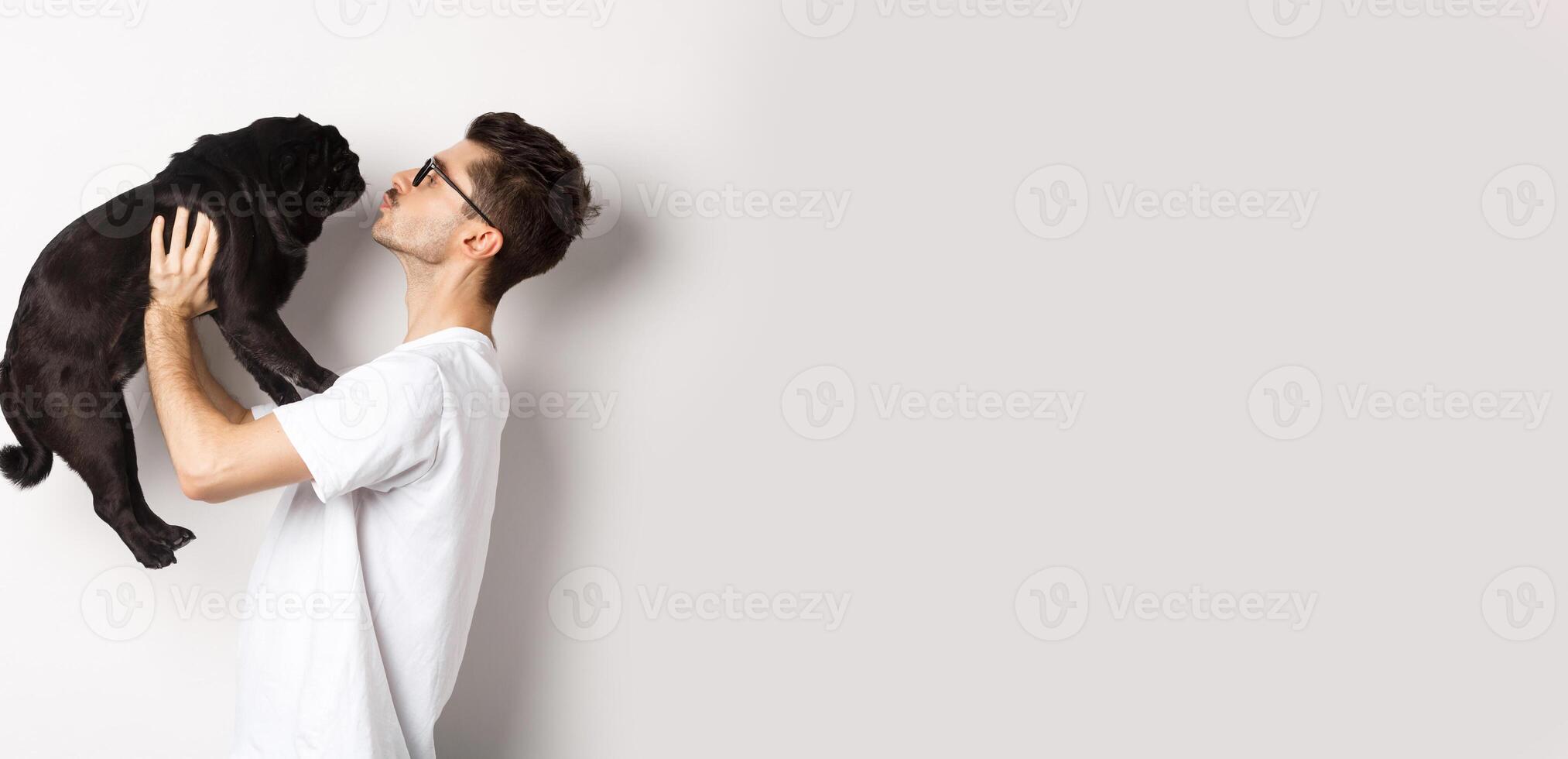 Profile of handsome young man kissing small cute dog face. Hipster guy loving his pug, standing over white background photo
