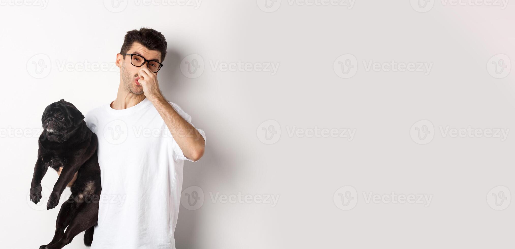Young man shut nose as holding pug, disgusted with bad smell fart of animal, standing over white background photo