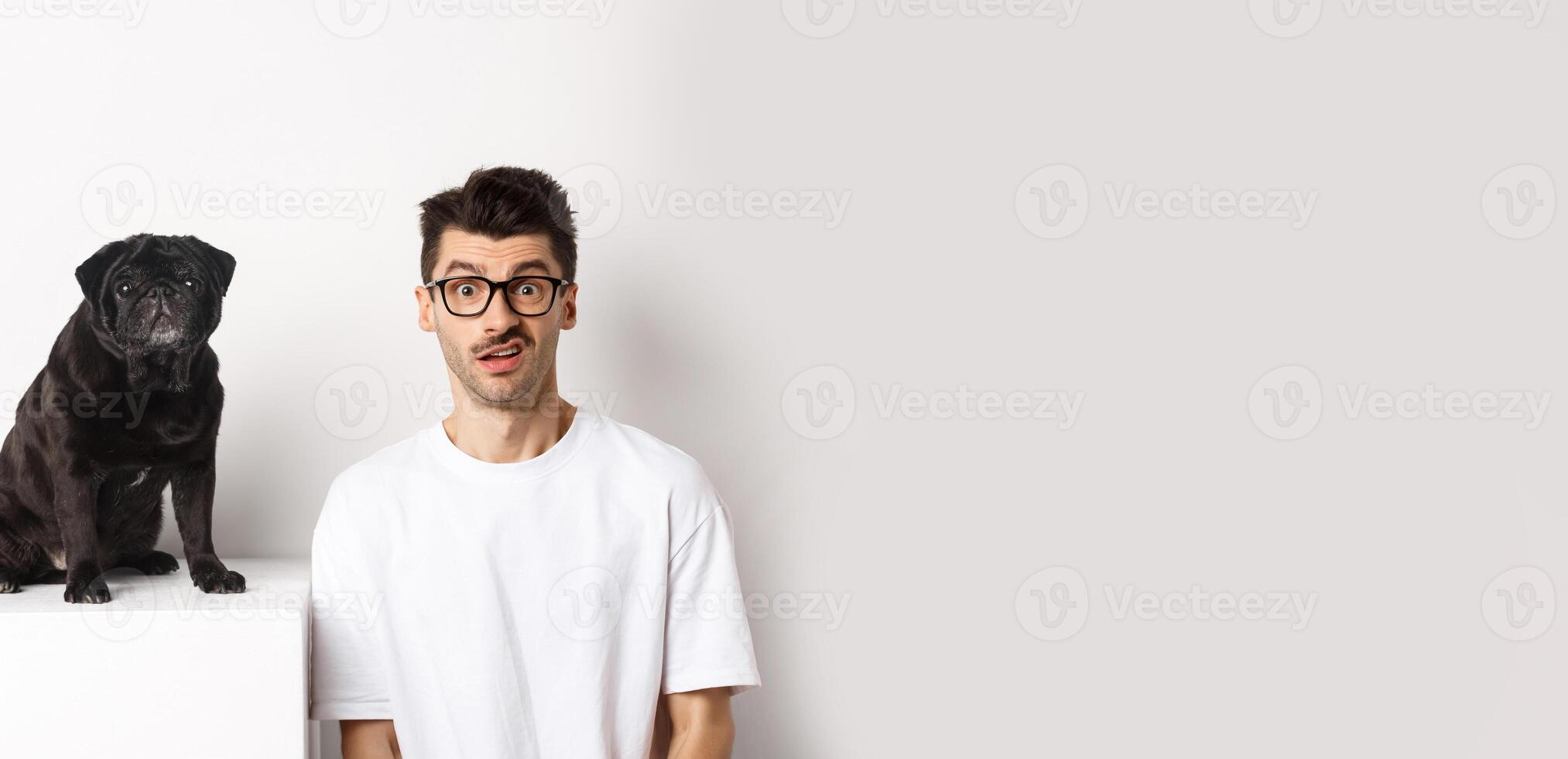 Image of hipster dog owner sitting near cute black pug, staring at camera confused and puzzled, white background photo