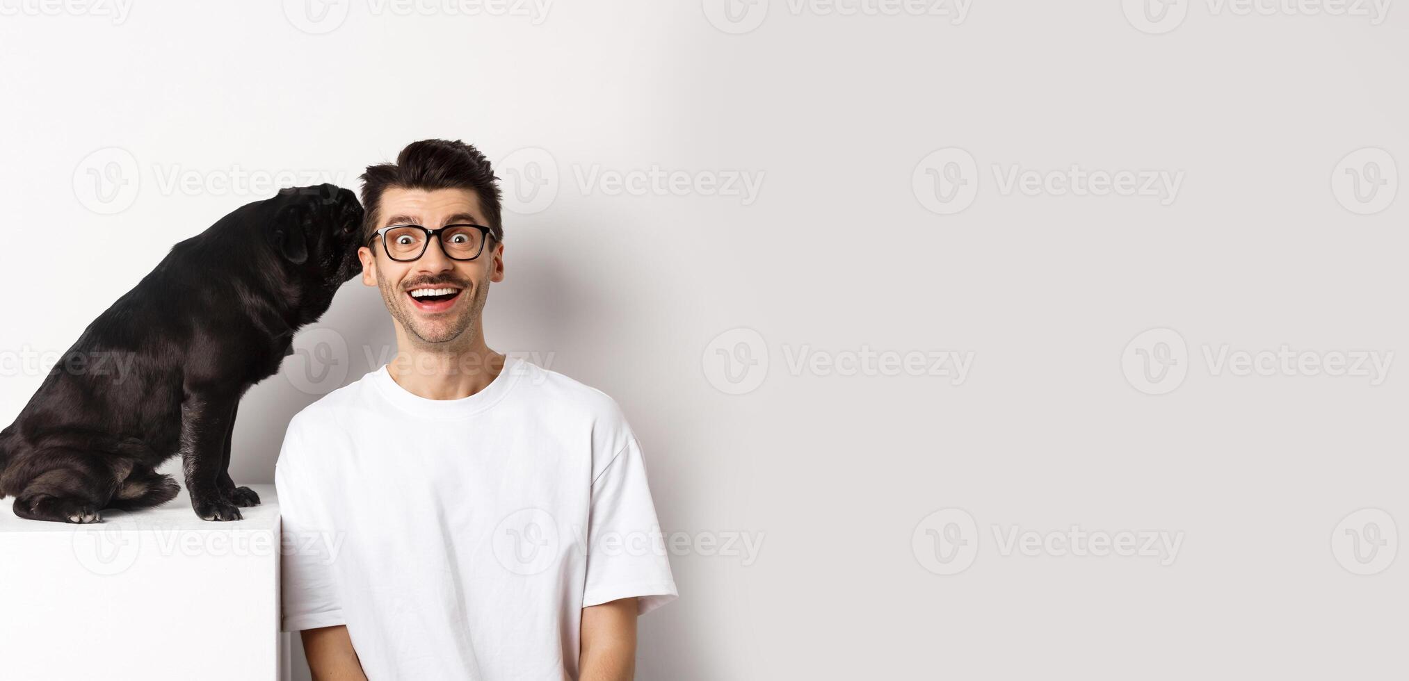 Image of cute black pug dog whispering at owner ear, man looking amazed and smiling, standing over white background photo