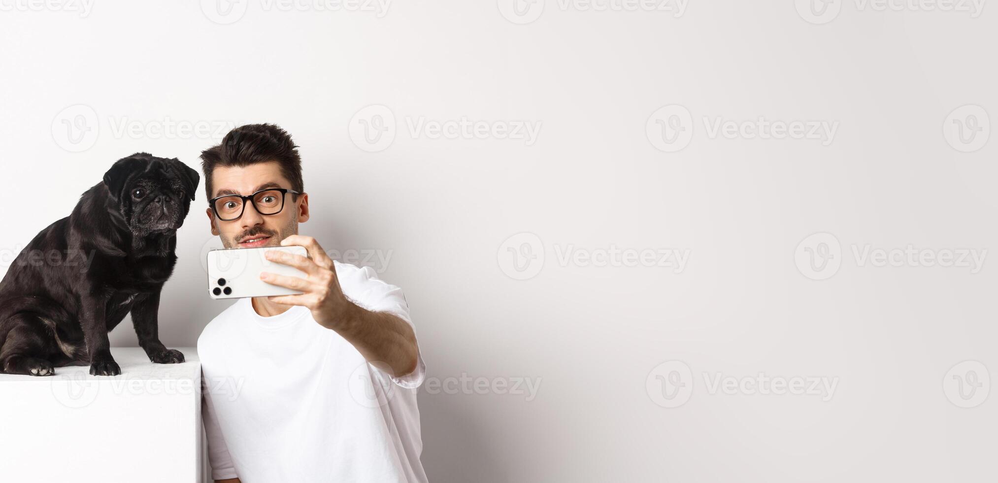 Image of handsome young man taking selfie with cute black dog on smartphone, posing with pug over white background photo