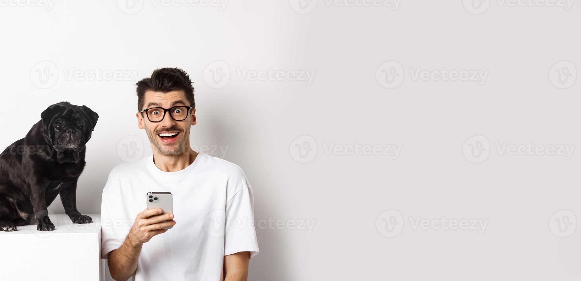 Cheerful young man hipster staring at camera, sitting with cute black pug dog and using mobile phone, standing over white background photo