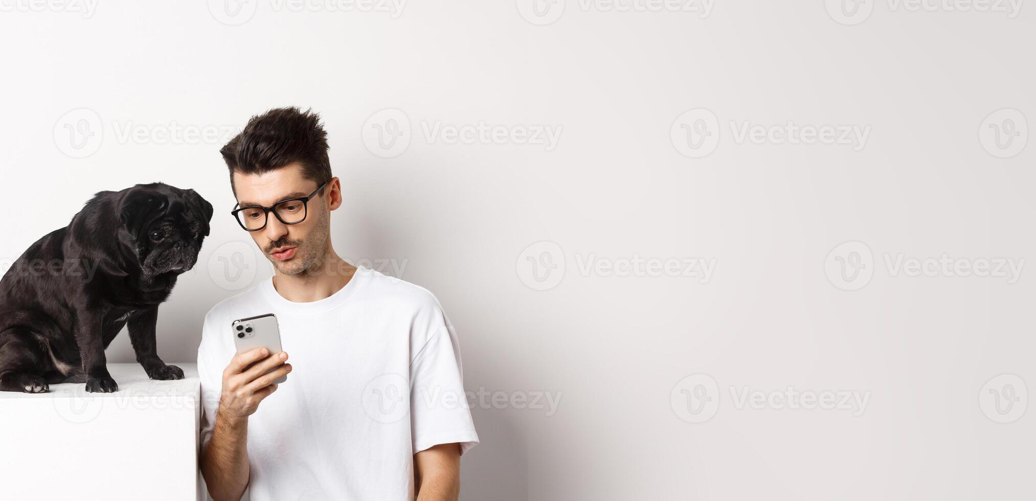 Young man showing photos on smartphone to cute dog, pet owner and pug staring at mobile phone, white background