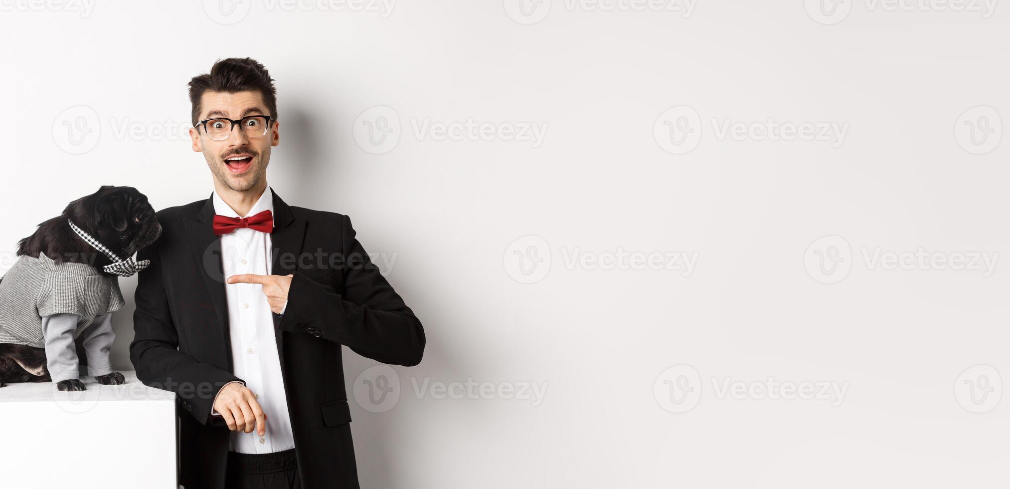Cheerful guy standing with his cute pug, pointing finger at dog wearing party costume, posing over white background photo