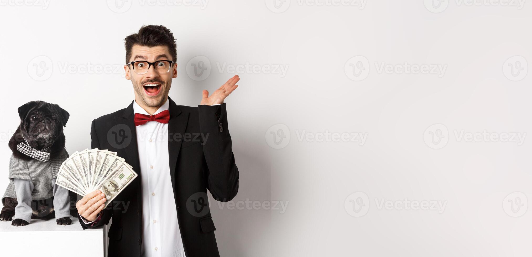 Happy young man and cute black dog standing in party costumes, pug owner holding money dollars, staring at camera amazed, white background photo