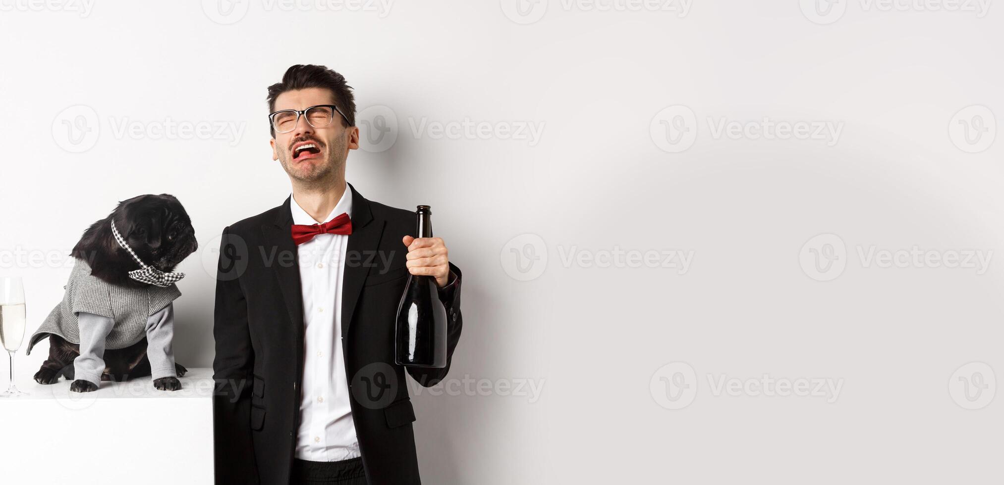 Sad man crying and drinking champagne from bottle while pug in cute party costume staring confused, standing over white background photo