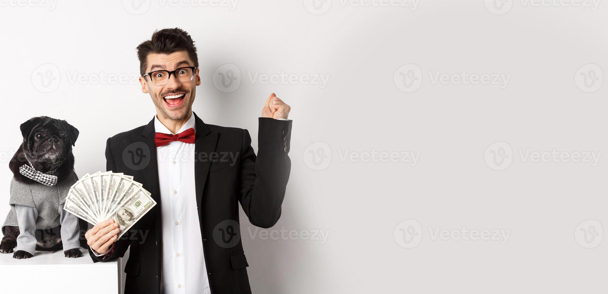 Happy young man in suit earn money with his dog. Guy rejoicing, holding dollars, black pug in costume staring at camera, white background photo
