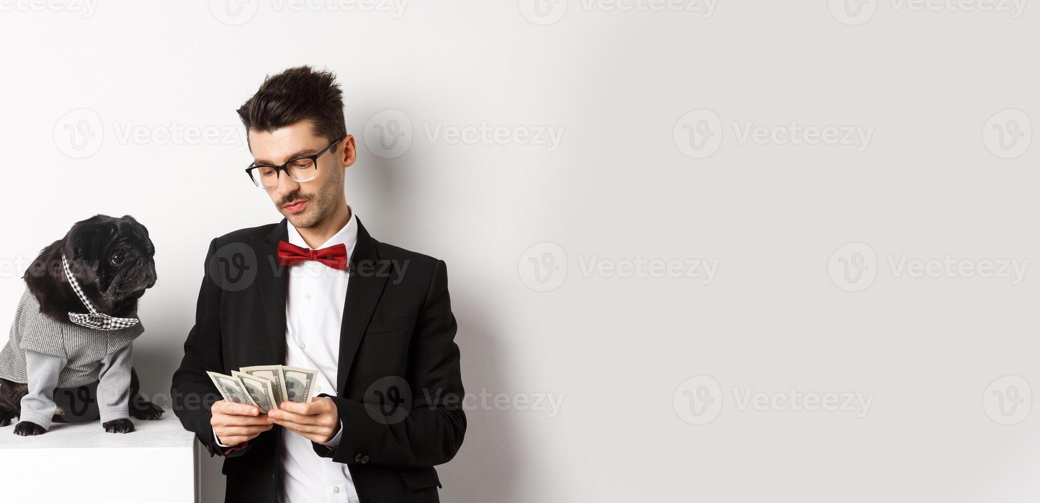 Handsome young man in suit standing near black pug in costume and counting money, working on parties, posing over white background photo