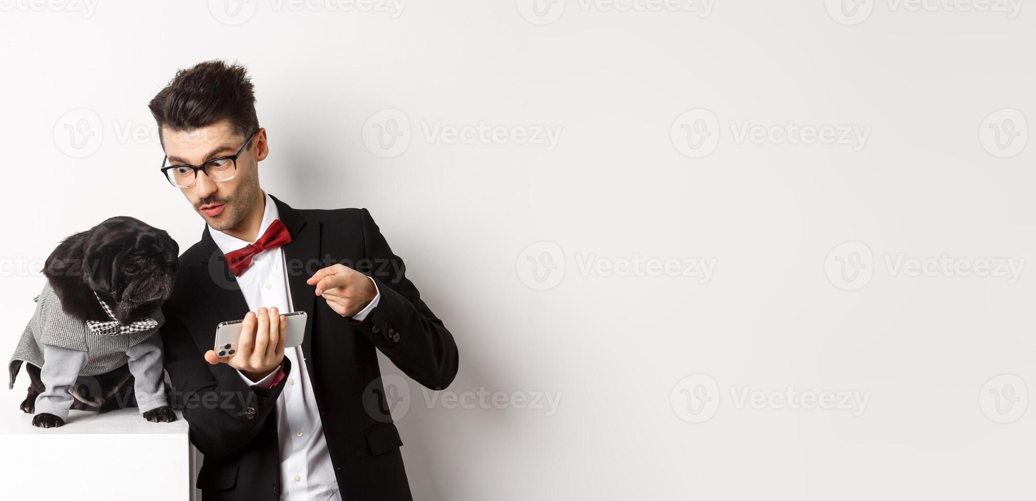 Handsome young man showing something on mobile phone to his dog. Owner shopping online with pet, standing in costumes over white background photo