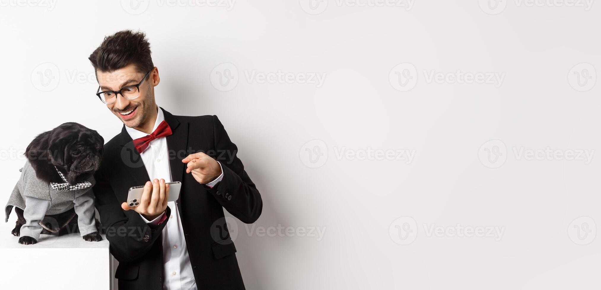 Handsome young man showing something on mobile phone to his dog. Owner shopping online with pet, standing in costumes over white background photo