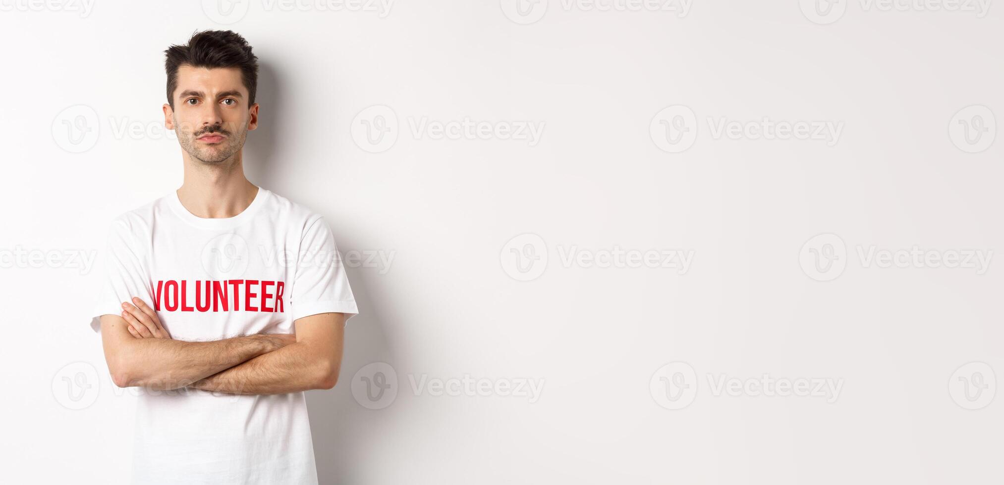 Serious young male volunteer in white t-shirt, holding arms crossed on chest, looking at camera, ready to help photo