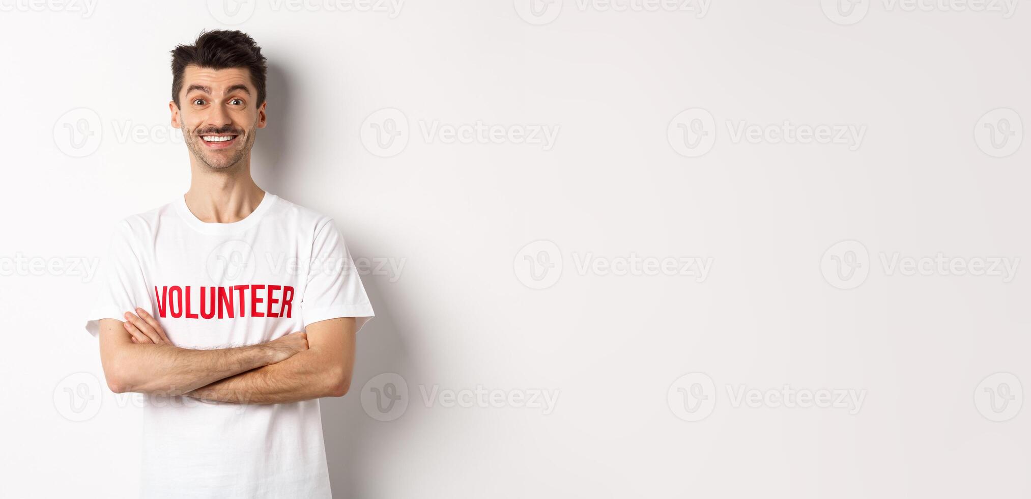 Happy young man in volunteer t-shirt ready to help, smiling at camera, cross arms on chest confident, white background photo