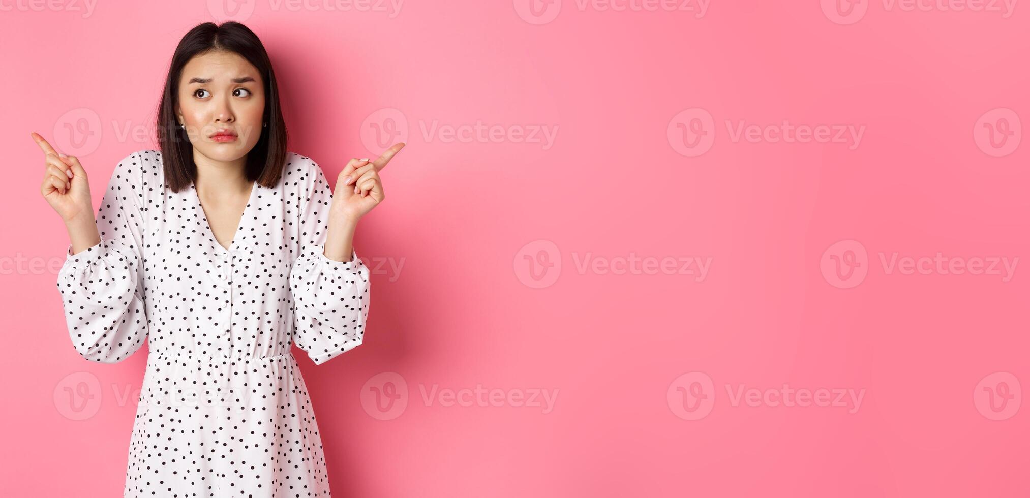 Confused asian woman hesitating, shrugging and pointing hands two sides, staring indecisive and need help with choice, standing over pink background photo