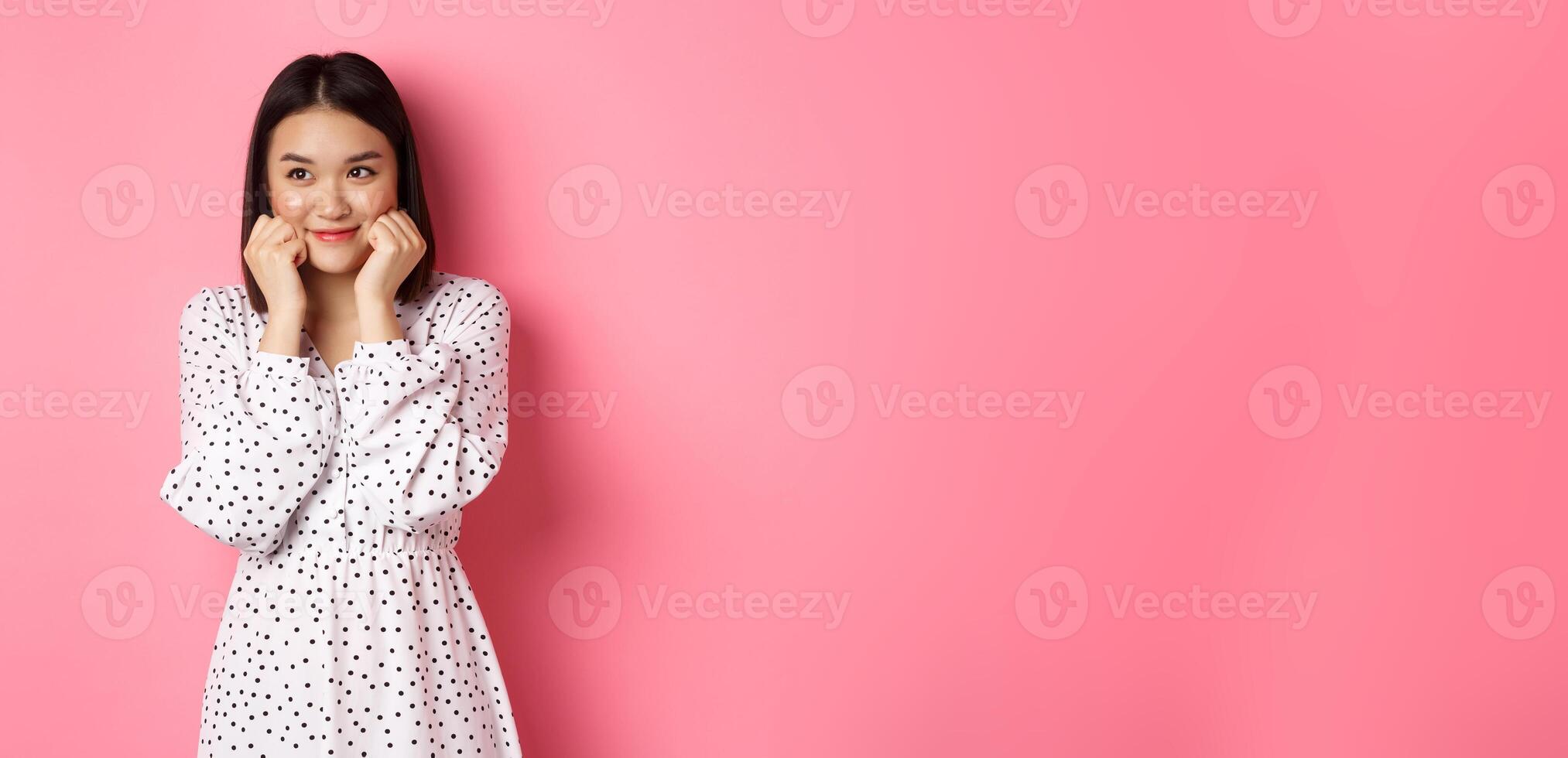 Cute and shy asian girl blushing, touching cheeks and looking left at copy space silly, standing against pink background photo