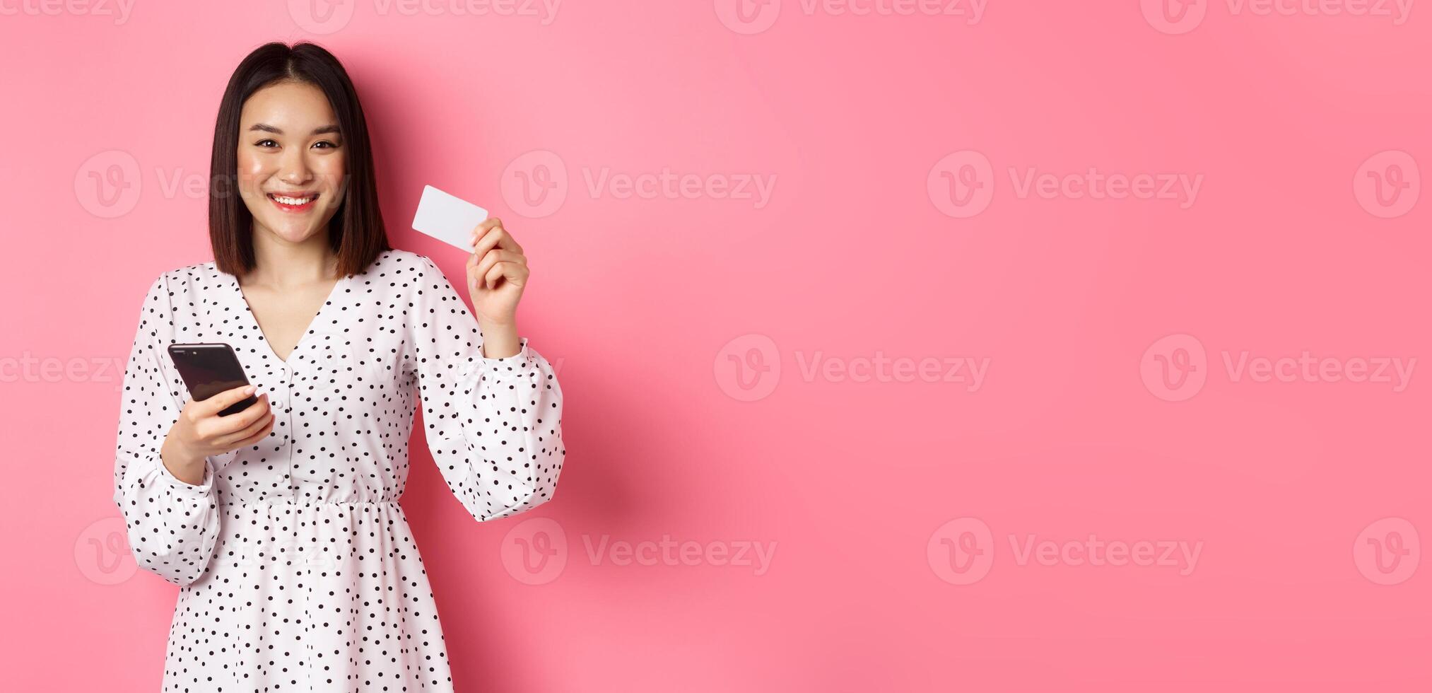 Attractive young asian woman order online, holding credit card and mobile phone, making internet purchase, standing happy over pink background photo