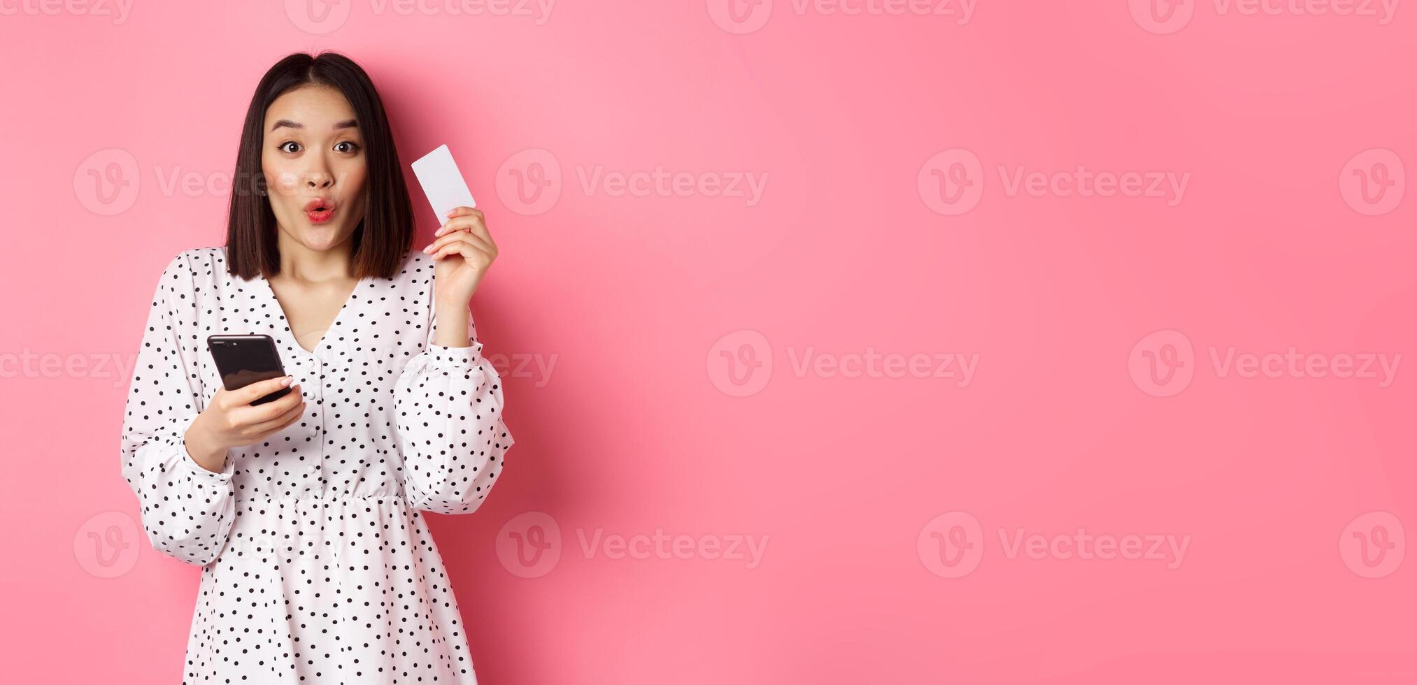 Attractive young asian woman order online, holding credit card and mobile phone, making internet purchase, standing happy over pink background photo