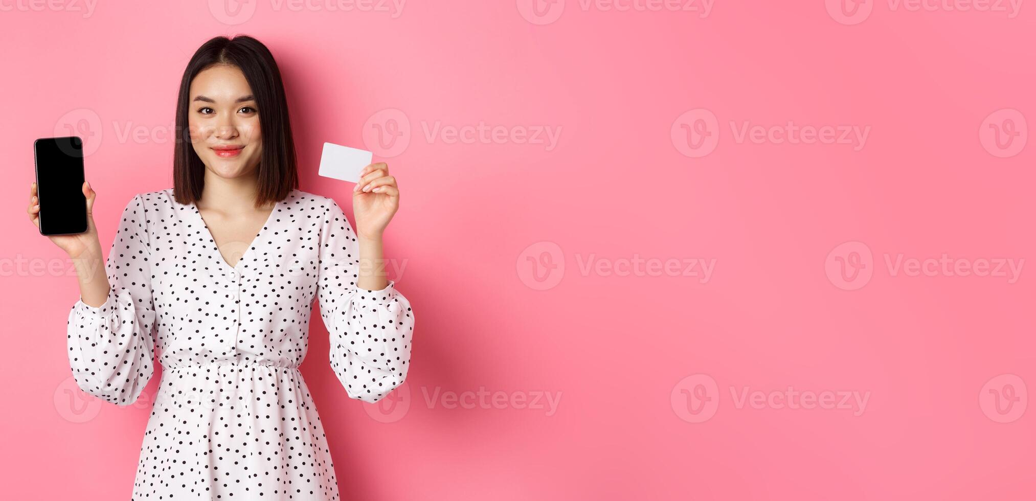 Cute asian woman shopping online, showing bank credit card and mobile screen, smiling and looking at camera, standing over pink background photo