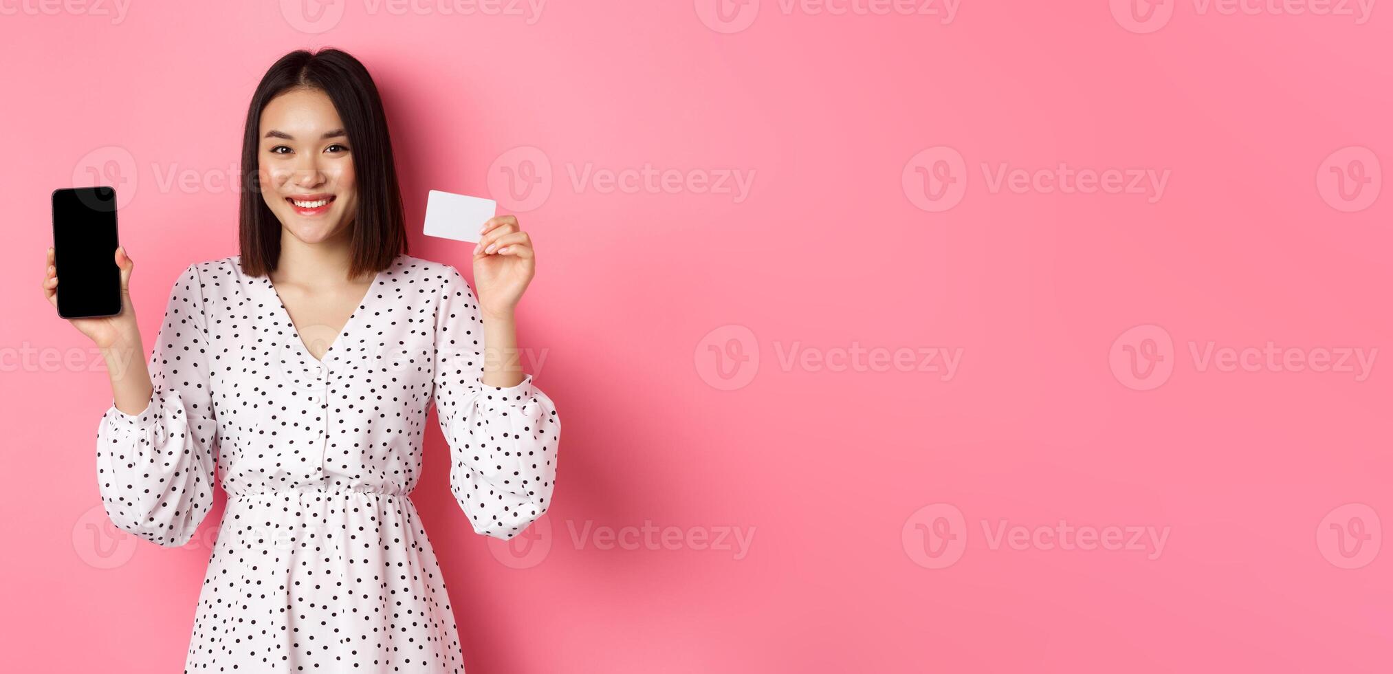Cute asian woman shopping online, showing bank credit card and mobile screen, smiling and looking at camera, standing over pink background photo