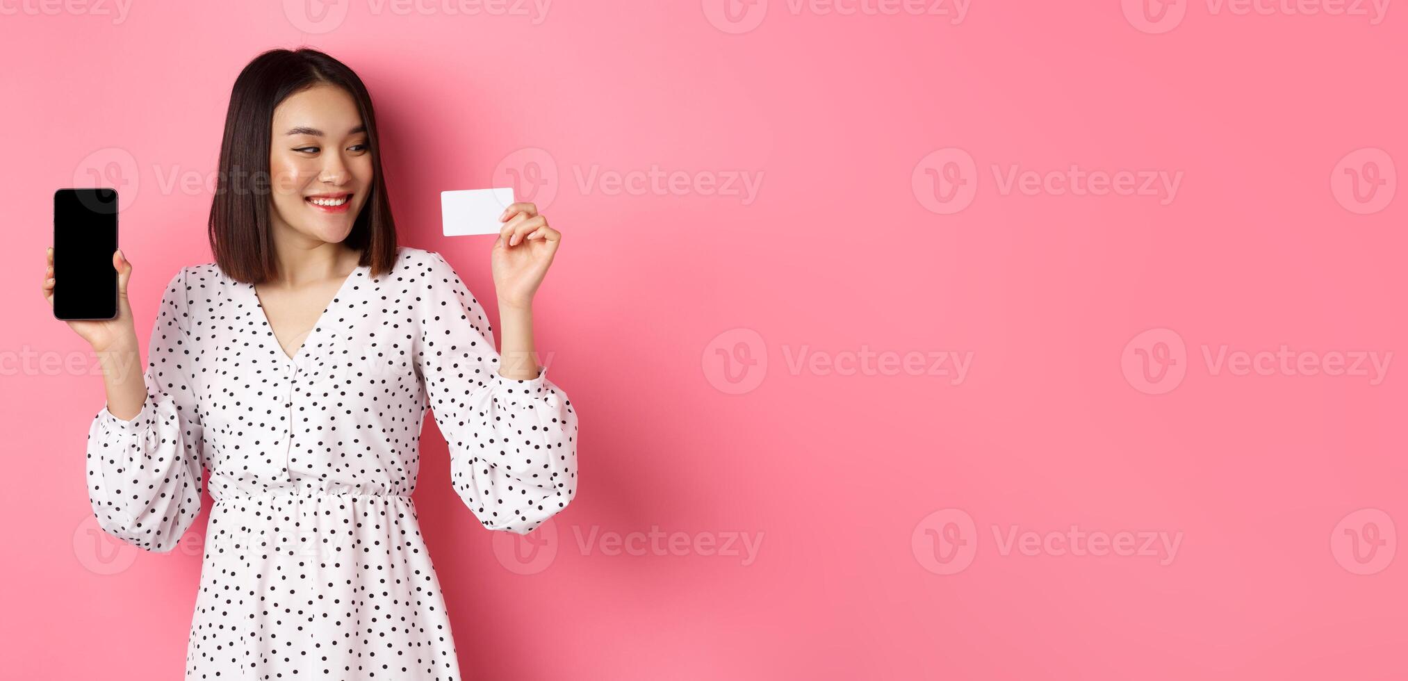Happy young asian woman internet shopping, showing smartphone screen and looking satisfied at credit card, standing over pink background photo