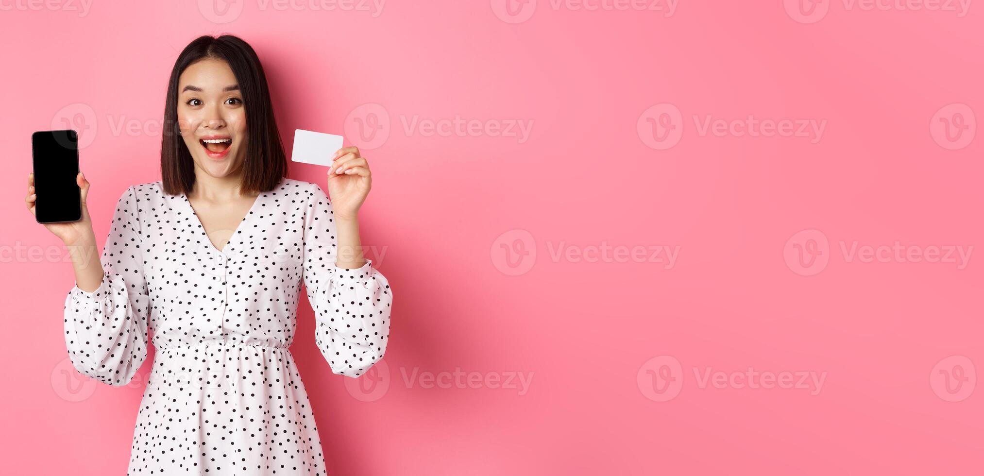Cute asian woman shopping online, showing bank credit card and mobile screen, smiling and looking at camera, standing over pink background photo