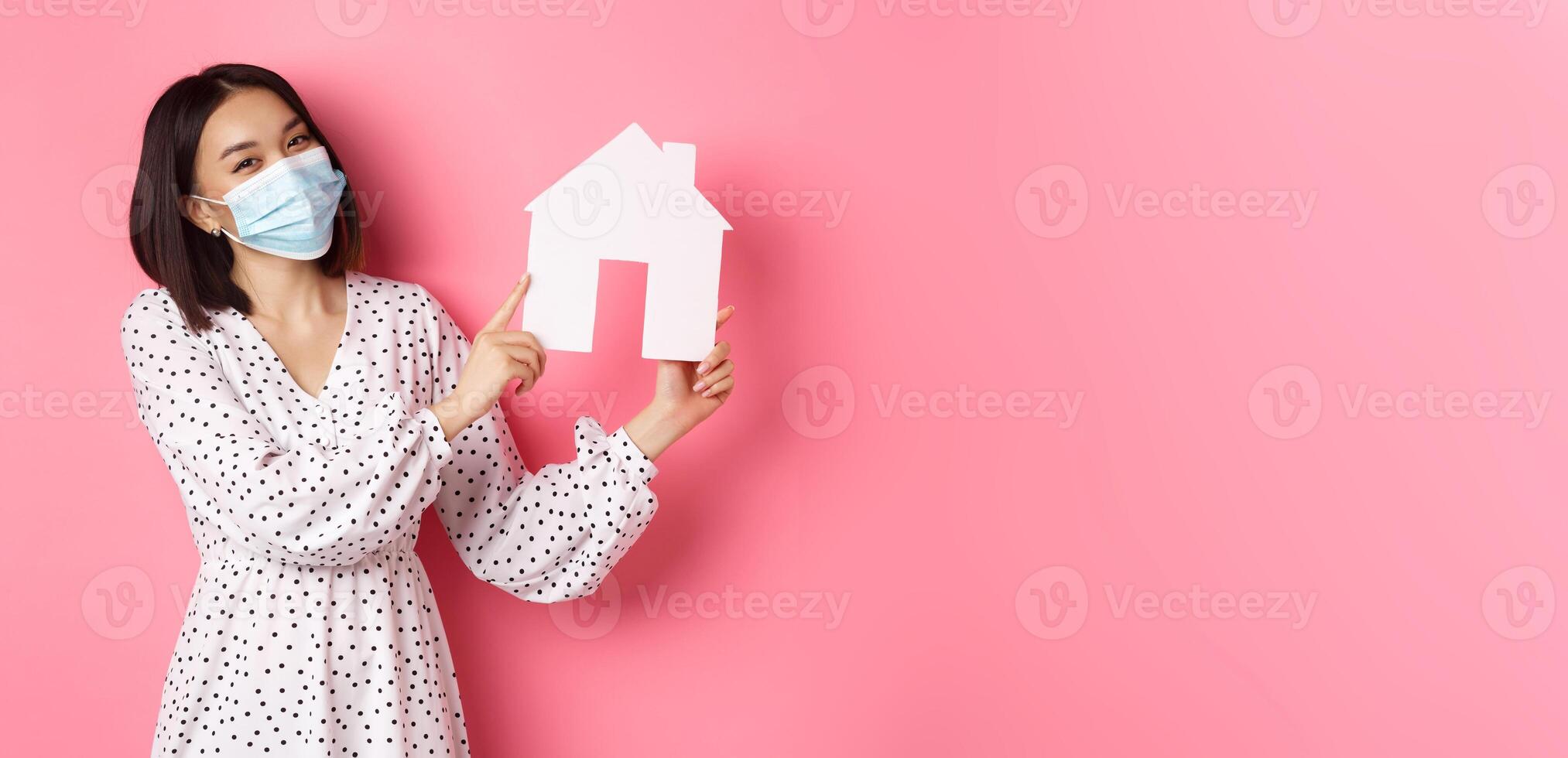 Covid-19, real estate and lifestyle concept. Cute asian woman in face mask selling houses, showing model of home and looking at camera, standing over pink background photo