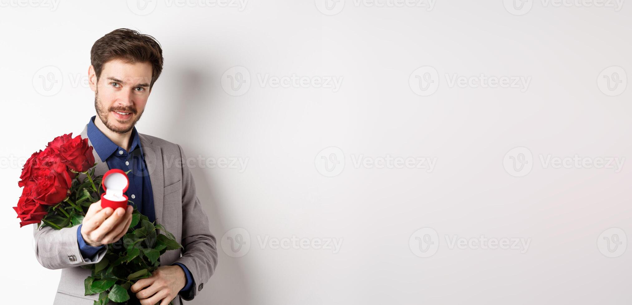 Romantic man with boquet of red roses asking to marry him, holding engagement ring and looking confident at camera, standing in suit over white background photo