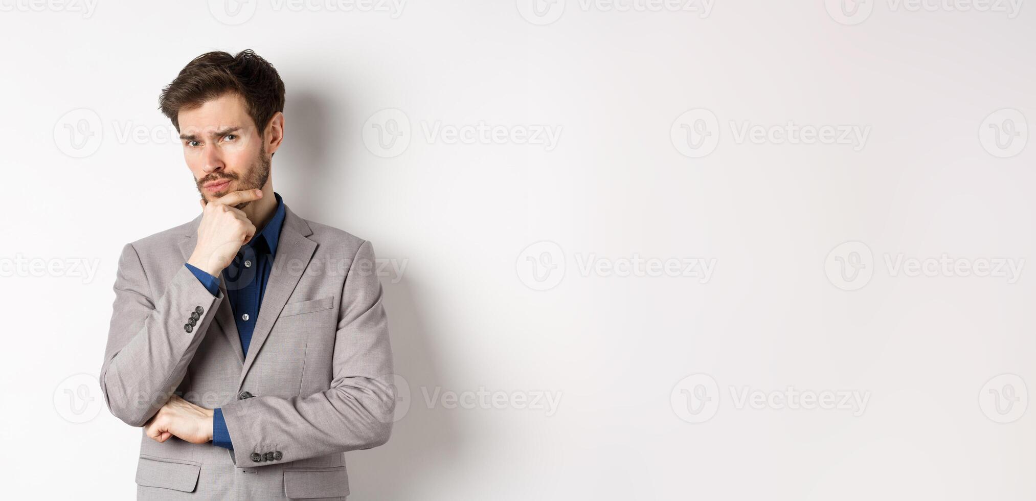 Hesitant young businessman in suit looking skeptical at camera, thinking and making decision, standing against white background photo