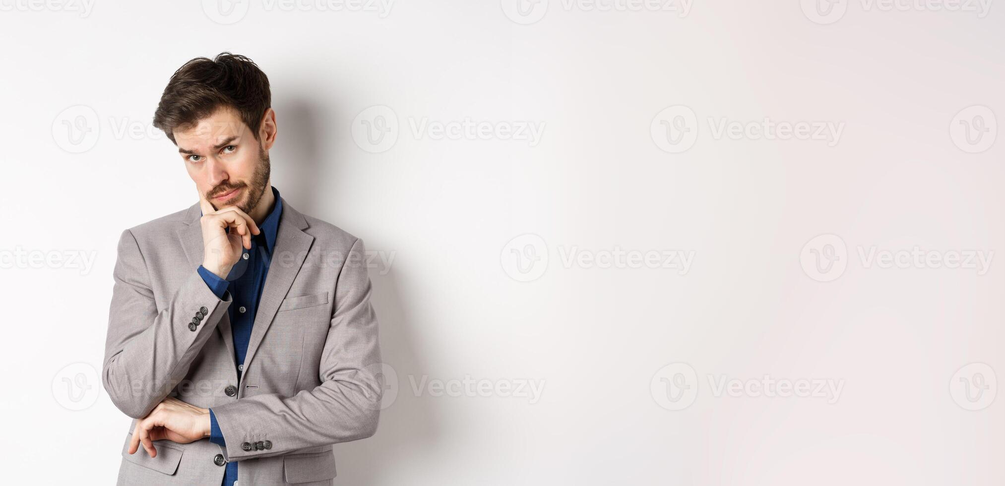 Hesitant businessman in suit making decision, looking pensive at camera with concerned face, having doubts, standing on white background photo