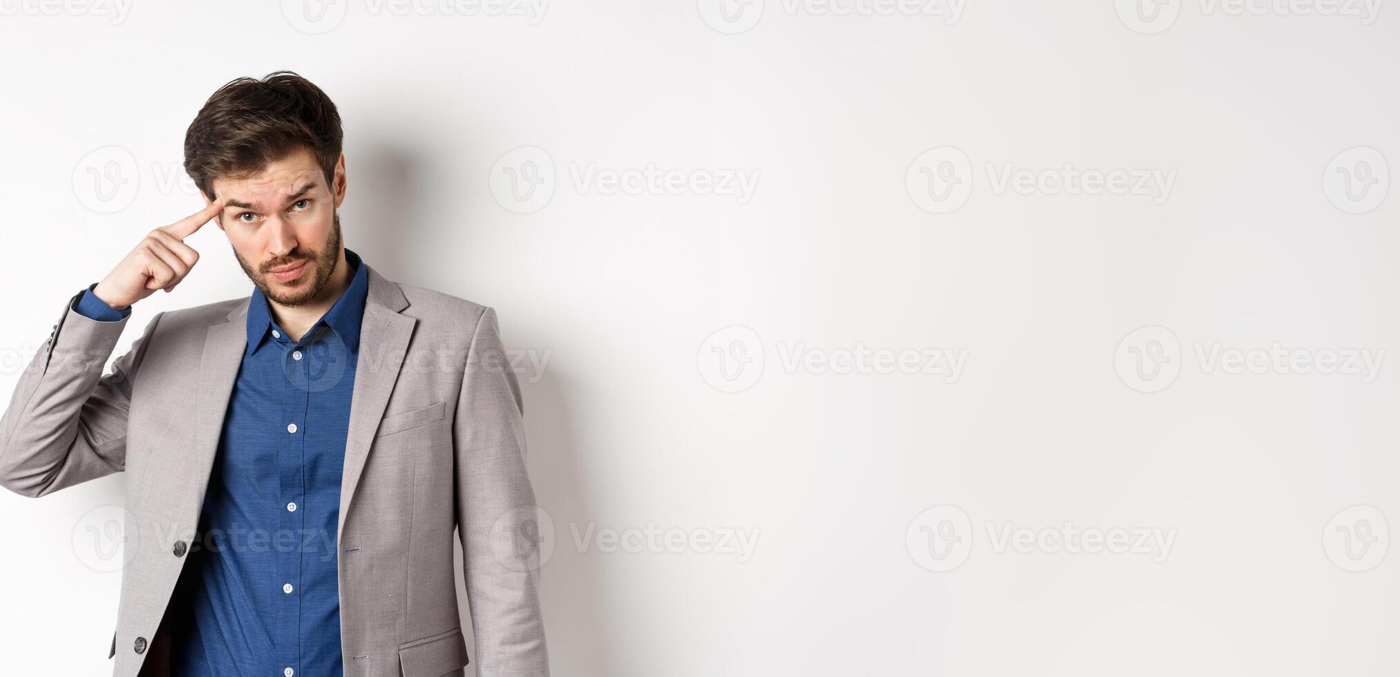 Annoyed male employer scolding personal, pointing finger at head, are you stupid out of your mind gesture, stare at camera from under forehead, standing in suit on white background photo